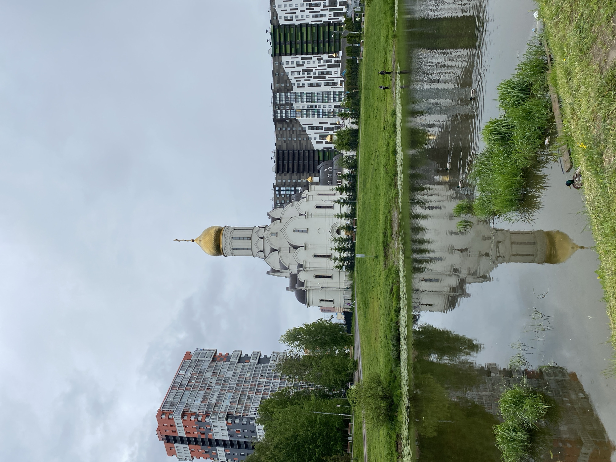 The image shows a church with a golden dome located in a city. The church is white and has several windows. There are modern buildings in the background and reflection of the church and buildings can be seen in the water. The location of the church and buildings appears to be a park or green space next to a river or lake. There are some people in the middleground walking near the bank, and a duck is visible in the water. The sky is overcast.