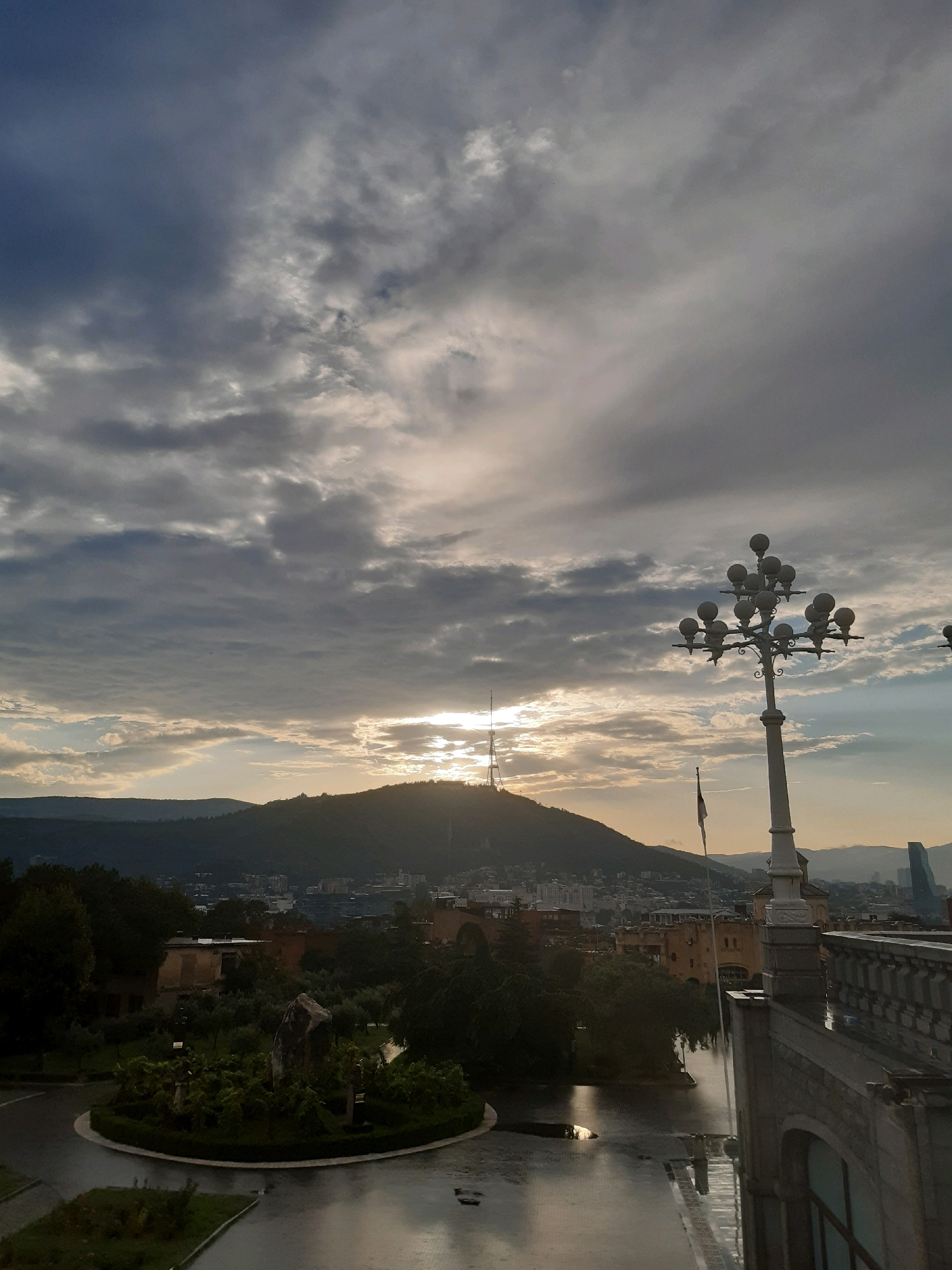 The image depicts a view of Tbilisi, Georgia, from a high vantage point, likely during late afternoon or early evening. The sky is filled with dramatic, cloudy formations, with the sun partially obscured by clouds, creating a soft, diffused light. In the distance, a prominent hill is silhouetted against the sky, with a television tower visible near its peak. The city stretches out below, with buildings of various architectural styles.
In the foreground, on the right side of the image, a tall, ornate lamppost with multiple globes stands prominently. Adjacent to it is a grey stone balustrade, suggesting the viewpoint is from a building with a balcony or terrace. To the left, a circular garden area is visible, featuring greenery and a large rock. The ground in front of the garden appears wet, indicating recent rain, and reflects the surrounding structures and the sky. A few scattered trees add to the landscape. In the far background, the silhouette of more distant mountains can be seen. There are no people or animals visible in the frame.
