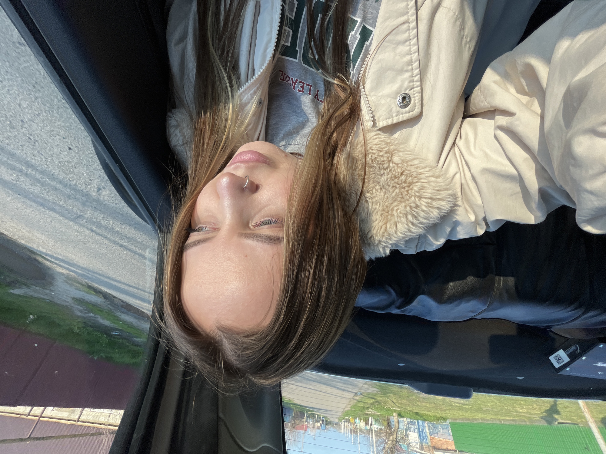 A medium shot captures a young woman inside a vehicle. She is wearing a light-colored jacket with a fur collar over a gray sweatshirt. The vehicle's interior is visible, including parts of the black dashboard and the car's window. The exterior is blurred, but suggests an urban environment, with buildings, fences, and foliage. The location is identified as Novopokrovskaya, Russia.
