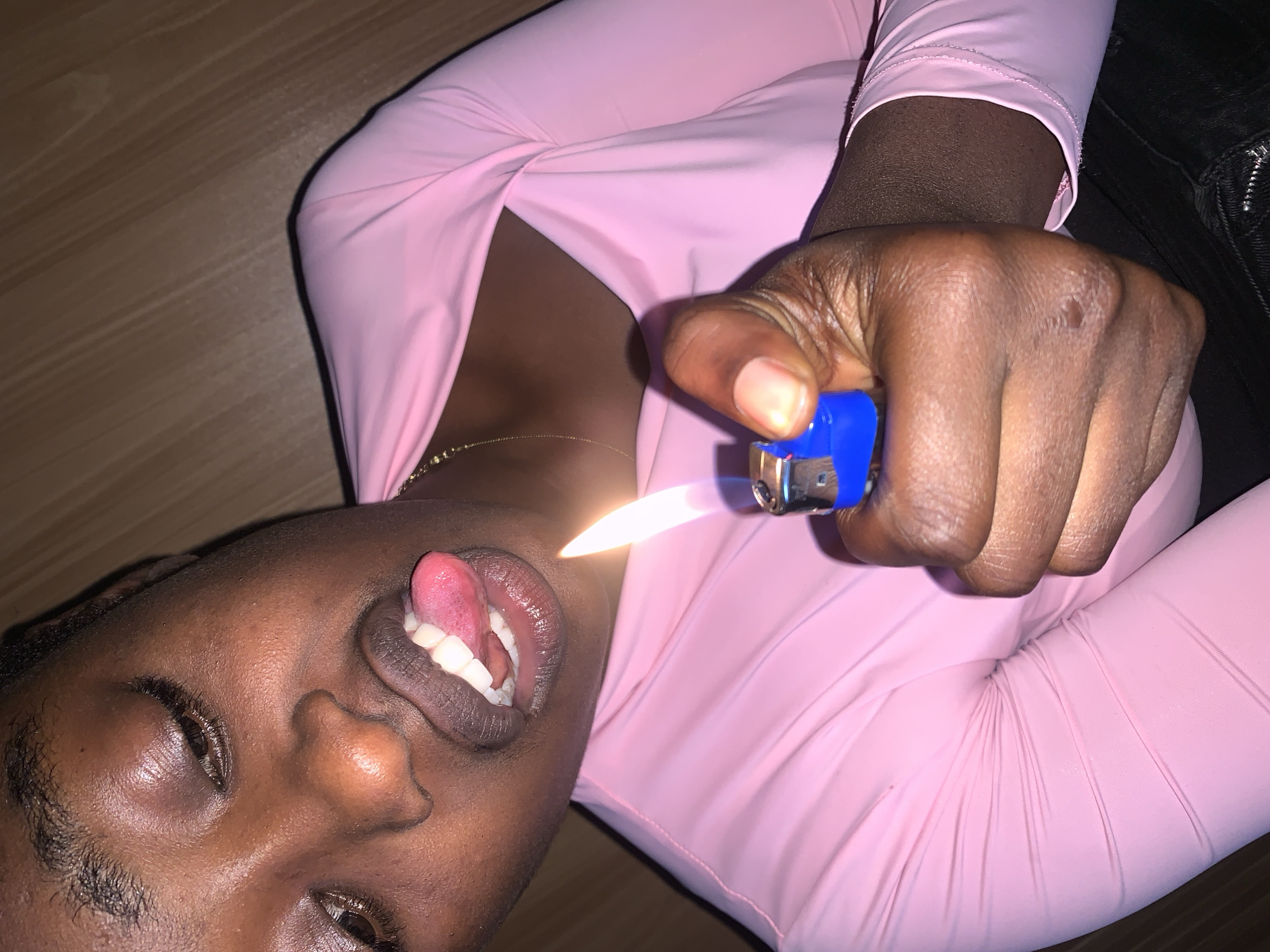 In Gura Top, Nigeria, a close-up shot captures a young woman with dark skin, her face tilted upwards. She wears a pink long-sleeved shirt. Her eyes are closed, and her mouth is slightly open, with her tongue extended. In her right hand, she holds a blue lighter that is lit, with a visible flame. The background is a wooden surface.