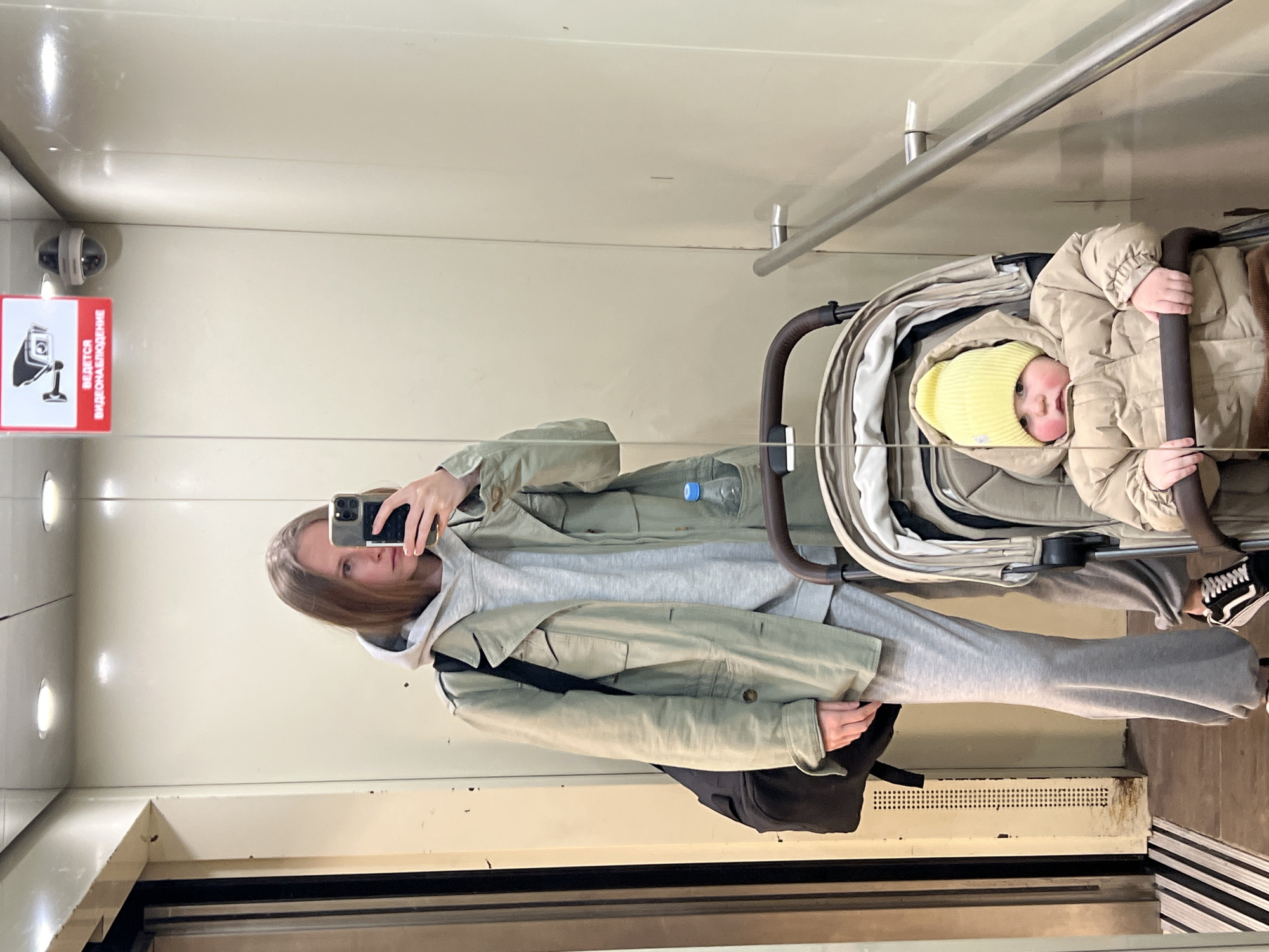 A person with medium-length hair and a light-colored jacket is taking a selfie in an elevator using a smartphone. The reflection shows the individual, who is wearing grey sweatpants and a hoodie underneath the jacket. A baby in a stroller, wearing a yellow hat and a beige puffy jacket, is also visible in the reflection. The elevator has plain, light-colored walls and a handrail. A surveillance camera and a sign in Russian indicating video surveillance are mounted on the wall near the top of the frame. A water bottle is visible in the stroller. The lower part of the elevator shows a vent and a striped floor. There is no indication of the location being Khimki, Russia.