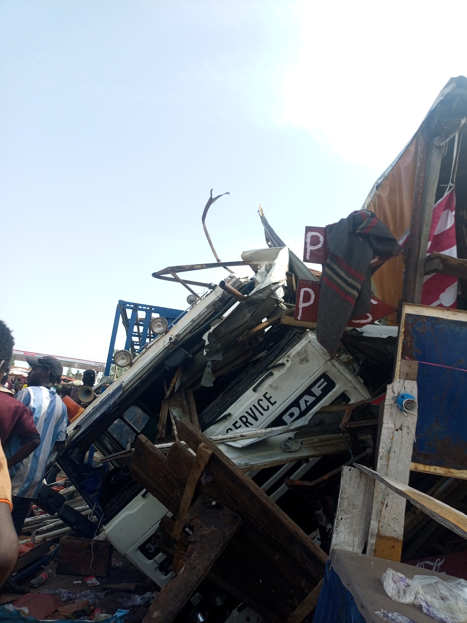 The image depicts a chaotic scene of debris and damaged structures in what appears to be the aftermath of an accident or collapse, in Gombe, Nigeria. A severely damaged white DAF truck, with the word "SERVICE" visible on its side, is buried under a pile of splintered wood, metal fragments, and other unidentified materials. The remnants of a structure, possibly a roadside stall or building, are visible, with a blanket and what appear to be parking signs hanging from the remaining framework. Several people are gathered around the wreckage, observing the scene. Some individuals appear to be wearing striped shirts. The background suggests an urban environment under a clear sky.