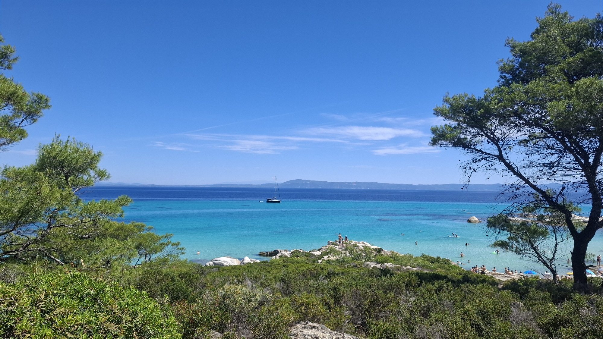 The image depicts a scenic coastal view in Toroni Municipal Unit, Greece, characterized by a clear blue sky, vibrant turquoise waters, and lush green vegetation. In the foreground, dense green bushes and rocky outcrops frame the scene, with pine trees on the left and right sides of the image contributing to the natural landscape.
The middle ground reveals a picturesque beach area with people engaging in various activities. Some individuals are sunbathing on the sand, while others are swimming or wading in the shallow, crystal-clear water. Several large, smooth white rocks are scattered along the shoreline, adding to the beach's unique topography. A few people are standing on a rocky promontory, seemingly enjoying the panoramic view.
Further out in the tranquil sea, a lone sailboat with a tall mast is visible, anchored or sailing slowly in the deeper blue waters. The horizon is marked by a distant, hazy shoreline, suggesting a wider expanse of land or mountains across the sea. The overall atmosphere is one of serenity and leisure, capturing a typical summer day at a Greek coastal destination.