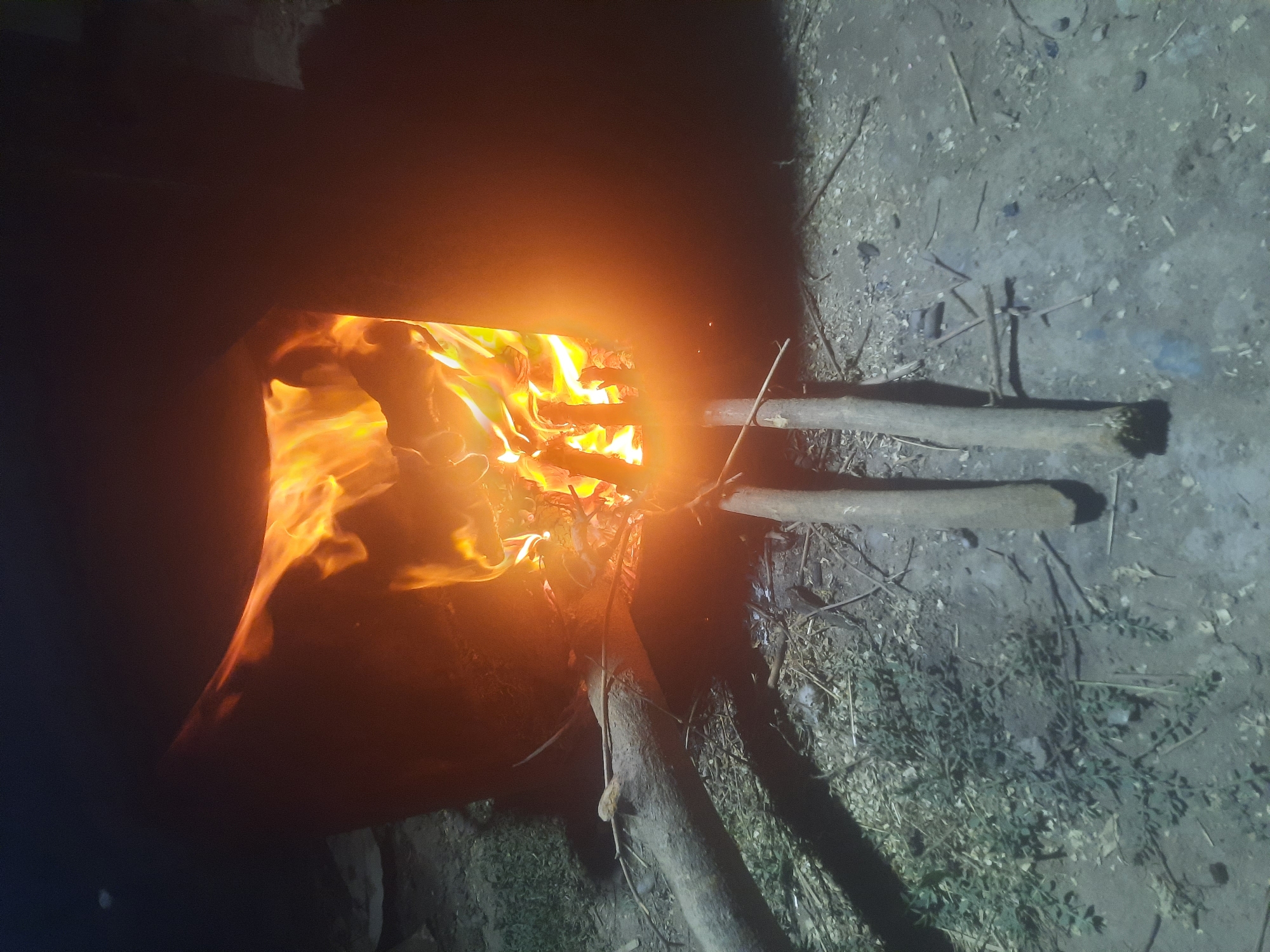 A nighttime outdoor scene depicts a rustic, metal fire pit containing a vibrant, roaring fire. The flames are intensely bright, casting an orange and yellow glow on the surrounding area. Two long, thin wooden sticks are placed in front of the fire pit, angled towards the flames. To the left of the fire pit, a thick, gnarled tree branch lies horizontally on the ground. The ground itself is covered in a mixture of dirt, small stones, and dried leaves. The overall impression is one of a simple, possibly rural, setting where a fire is being actively maintained for warmth or cooking.