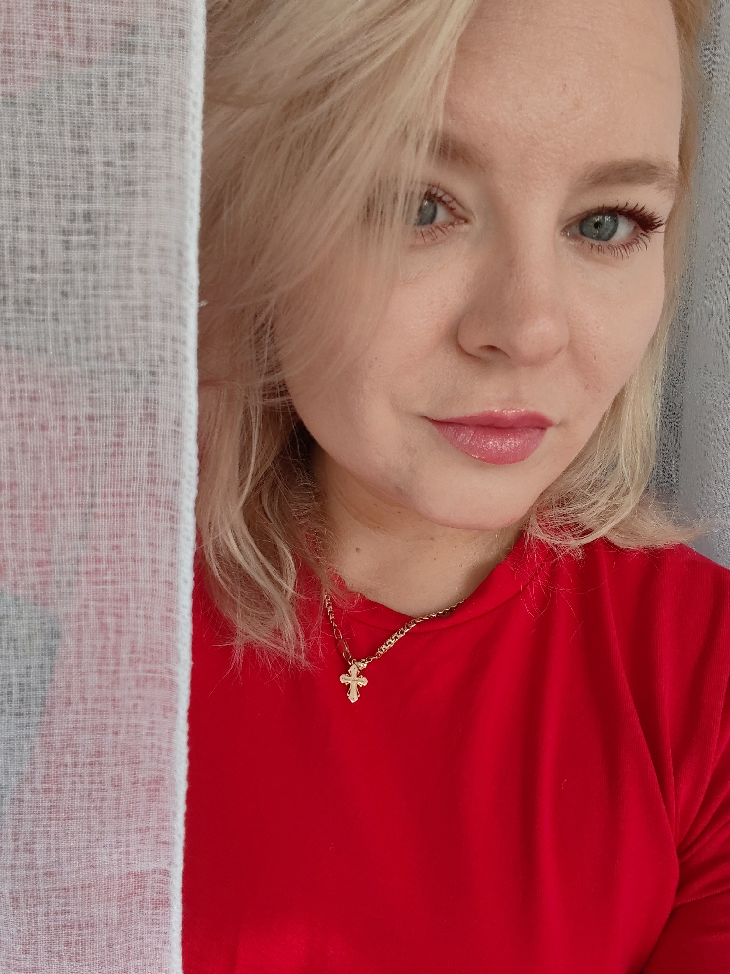 A medium shot portrait of a blonde woman with blue eyes, wearing a red t-shirt and a gold chain with a cross pendant. She is partially obscured by a sheer white curtain on the left side of the frame. The woman is looking towards the camera with a slight smile. The background is out of focus. The lighting appears to be soft and natural. The scene is indoors.