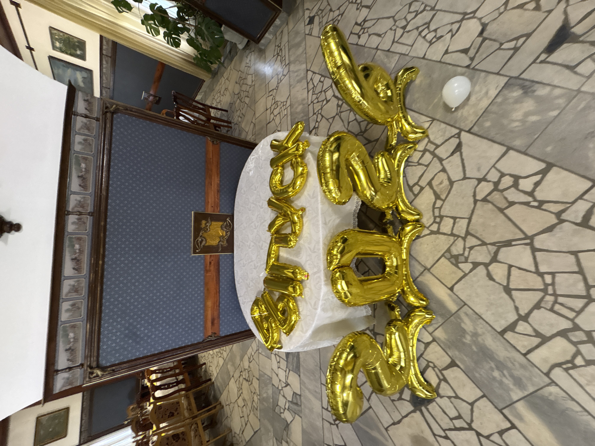 The image shows a table covered with a white cloth, on which gold letter balloons spell out "HAPPY BIRTHDAY". The balloons are arranged in two lines. The first line, on top, reads "HAPPY" and the second line, below, reads "BIRTHDAY". The table is set on a stone floor with irregular grey and beige tiles. In the background, there is a large, ornate wooden structure with blue patterned fabric. Several chairs and a plant are visible. To the right of the balloons, a single white balloon lies on the floor. The scene appears to be indoors, possibly in a hall or a room with traditional decor. The lighting suggests it could be daytime, but the exact time is not discernible. There are no people visible in the image.