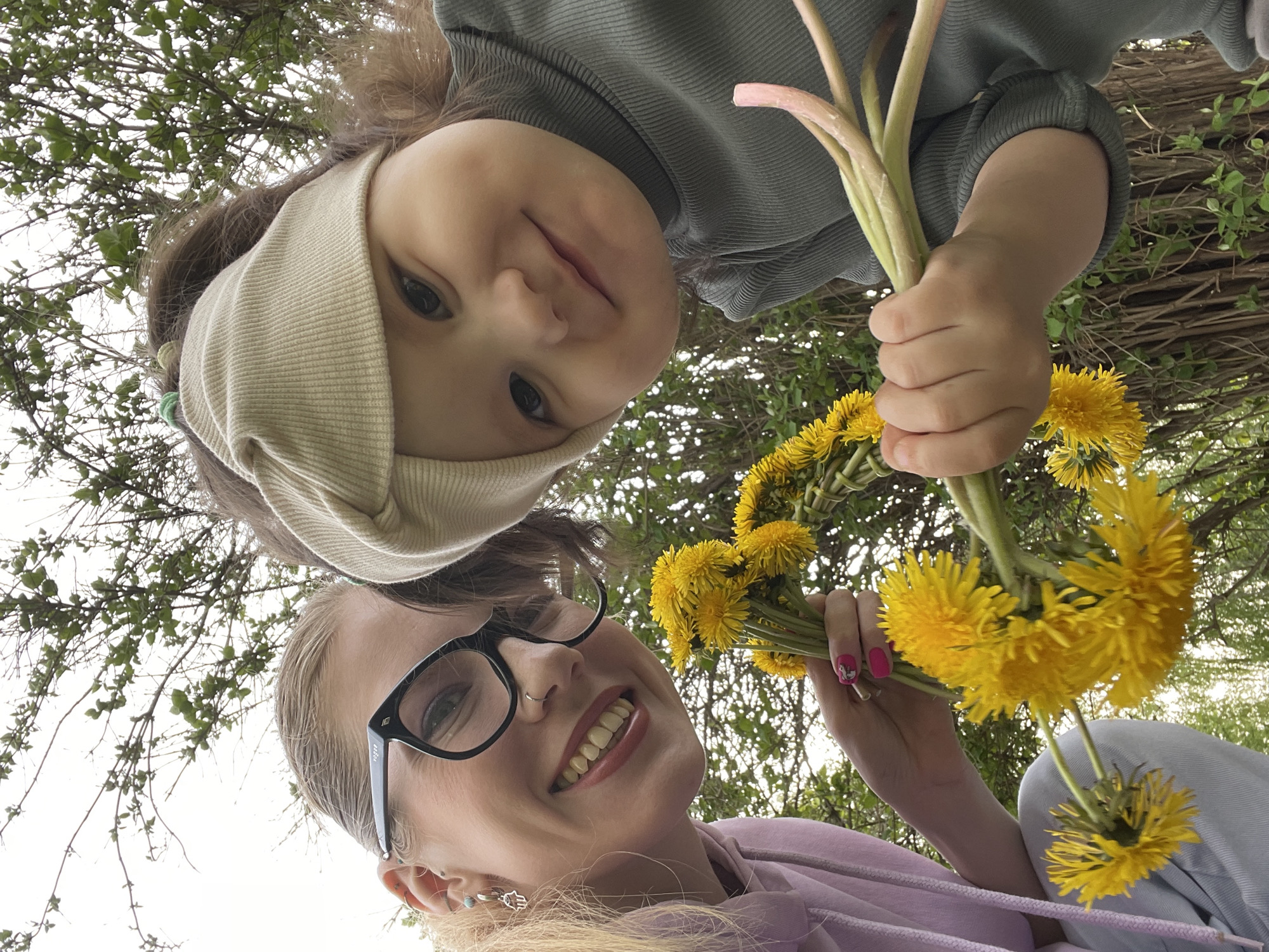 A low-angle shot depicts a woman and a child, both smiling, against a backdrop of tree branches and a cloudy sky. The woman, wearing black-framed glasses and a lavender-colored hoodie, holds a bouquet of bright yellow dandelions. The child, positioned above the woman, wears a beige headband and a muted green shirt, clutching what appears to be a collection of green stems. The composition suggests they are outdoors. Location is unspecified but could be in Izhevsk, Russia.