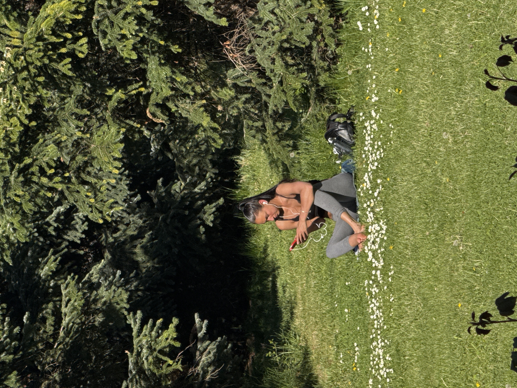 A woman is lying on her side in a grassy area next to a coniferous tree. She is wearing gray leggings and a black sports bra. She is using a smartphone with headphones plugged in. A backpack is to her right. The grass has many small white flowers growing in it. There are shadows on the grass from nearby vegetation. The location is in Sofia, Bulgaria.