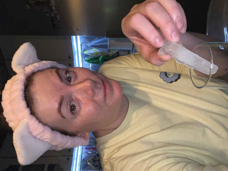 A woman with a pink cat-ear headband holds a large, clear ice chunk over a glass. She wears a light yellow t-shirt with a flower emblem. Her fair complexion is visible. The background reveals kitchen cabinets with a decorative lizard figure and a lit-up space with various items. The image is slightly tilted.