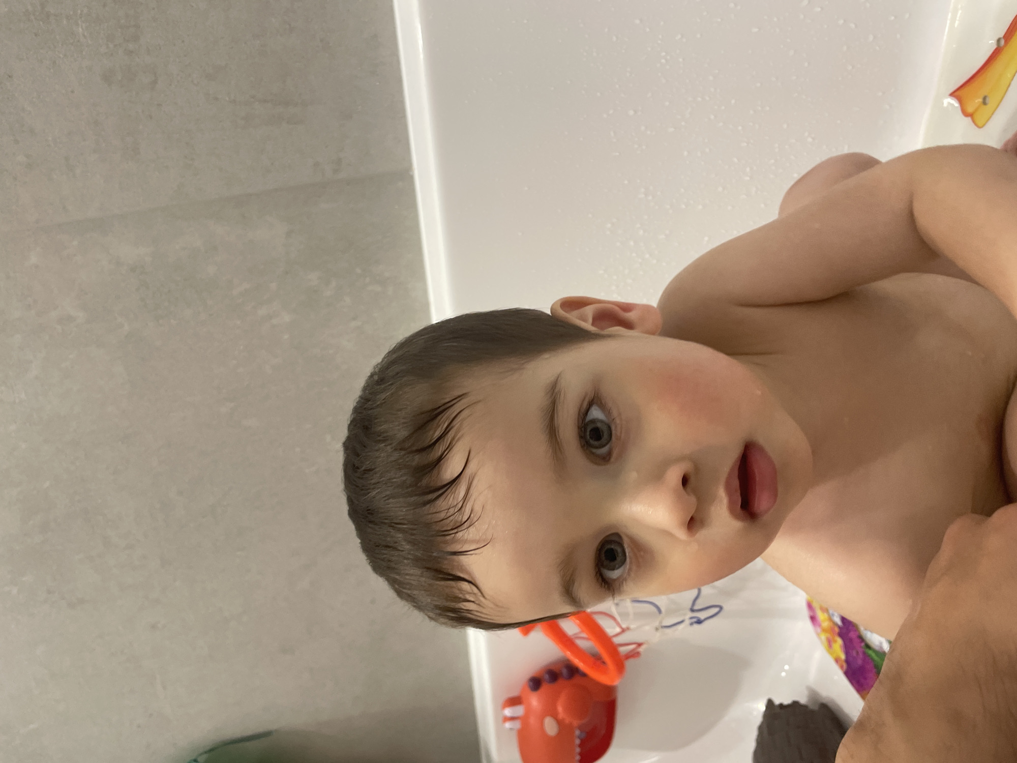A young child, likely a toddler, is partially visible in a bathtub filled with water. The child has wet, dark hair and is looking up at the camera with their mouth slightly open. The child's skin is fair, and their cheeks are flushed.
Several bath toys are visible in the tub, including a red toy resembling a dinosaur and a brightly colored object near the edge of the tub. The bathtub appears to be white and is set against a wall.
It is not possible to discern the location as Izhevsk, Russia from the given image. The scene is generally domestic and typical of a child's bath time.