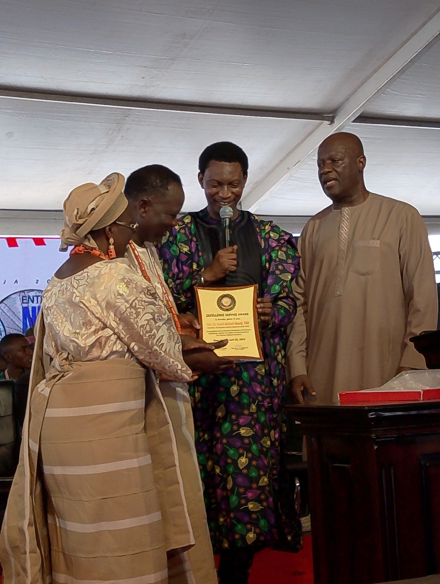Three individuals are centrally featured in an indoor setting. A woman, adorned in a traditional ensemble including a headwrap, ornate earrings, and a layered dress with horizontal stripes, stands to the left. A man in a patterned, multicolored robe holds a framed "Excellence Service Award" while speaking into a microphone. To the right, a man in a plain, light brown tunic looks on. The background includes a large screen with partial text visible, and the setting appears to be under a tent-like structure. A dark wooden table is situated on the right, with a red box on top. The image's location includes Dushepe, Nigeria, according to the award.