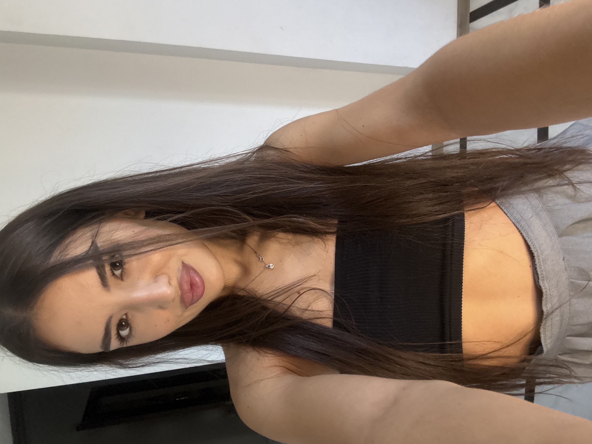 A young woman with long, dark hair and brown eyes is taking a selfie in a well-lit room. She is wearing a black strapless top and grey pleated skirt. A delicate necklace with a teardrop pendant is visible around her neck. The background shows a plain white wall and a dark doorway on the left, with a patterned floor visible at the bottom right. The lighting suggests an indoor setting, possibly a residence or studio. There is no specific indication of the location being in Chișinău, Moldova, or any other city.
