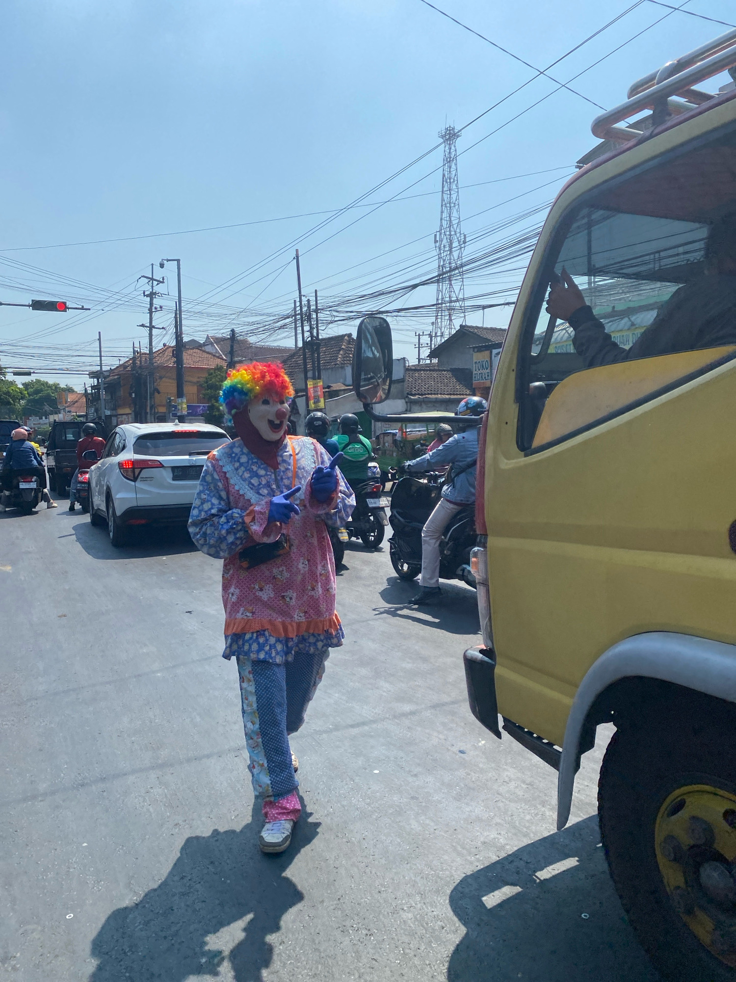A person dressed as a clown with a rainbow-colored wig, white face paint, and a colorful costume is walking on a street in Kediri City, Indonesia. The clown is wearing a pink and blue outfit with matching pants and white shoes. They are holding blue gloves and appear to be interacting with traffic. Several vehicles, including cars and motorcycles, are visible on the street. The environment appears to be an urban setting with buildings, power lines, and traffic signals. The sun is shining, casting shadows on the road.
