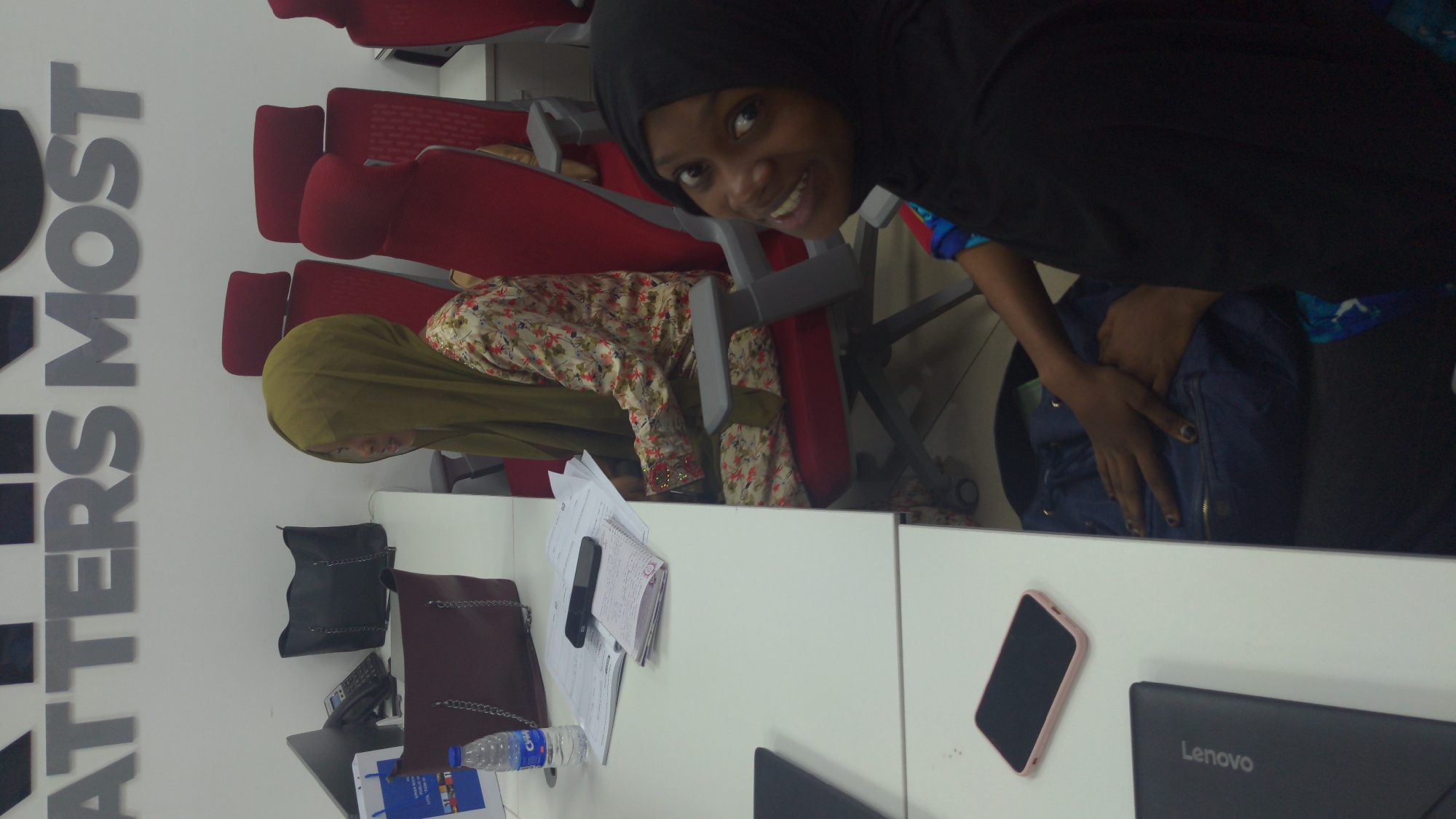 This image depicts an indoor scene, likely an office or meeting room, characterized by white tables and numerous red chairs with gray frames. Large, gray three-dimensional letters spelling "ATTERS MOST" are prominently displayed on a white wall to the left.
In the foreground, a young woman wearing a black hijab and a colorful patterned shirt smiles warmly as she looks directly at the camera. Her right arm is visible, resting on a blue, padded bag positioned next to her. Behind her, another woman is seated in a red chair, facing away from the camera. She is dressed in a floral long-sleeved top with embellished cuffs and a light olive green hijab. A third person, also in a similar floral top, is partially visible further in the background.
The white tables in the foreground are arranged with various personal and work items. On one table, there are two handbags—one black and one dark brown with a chain strap—alongside a clear plastic water bottle with a blue "Chi" label. A stack of papers and a black feature phone are also present. On an adjacent table, a pink-cased smartphone and a black laptop bearing the "Lenovo" logo are visible.
The scene is brightly lit by indoor lighting. The smiling expression of the woman looking at the camera conveys a positive and inviting atmosphere.