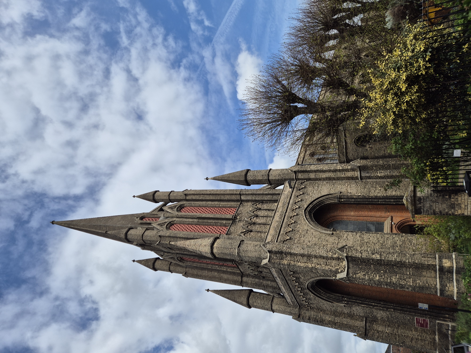 The image displays a towering, historic church building under a bright, partially cloudy blue sky. The church, likely of Victorian Gothic Revival architecture, features intricate brickwork in shades of grey and dark beige, accented with red brick infill within its arched openings and pointed windows. A prominent, ornate spire dominates the left side of the structure, flanked by smaller pinnacles.
On the right side of the image, several tall deciduous trees, mostly bare-branched, stand against the sky, some showing early signs of yellow-green foliage. A dense bush with variegated yellow and green leaves is positioned in front of a black metal fence, which runs along the base of the trees and separates the church grounds from what appears to be a public area.
The sky is a clear blue, adorned with scattered white cumulus clouds and a distinct, thin contrail left by an aircraft. The time of day appears to be bright daytime, likely morning or early afternoon, with ample natural light illuminating the scene. No people are visible, and the scene is static, capturing the architectural beauty and natural surroundings.
Several signs are visible near the fence and church wall, but their text is illegible due to distance and partial obstruction. The overall impression is one of a well-maintained, traditional church building set within a green, urban or semi-urban environment, consistent with locations in West Suffolk, United Kingdom. The image is taken from a low-angle perspective, looking upwards at the church and sky.