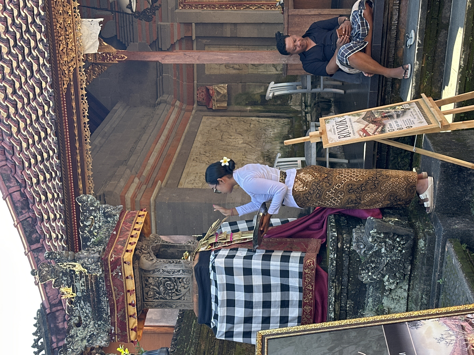 A medium shot captures a Balinese woman in traditional attire, a white long-sleeved blouse and a patterned brown sarong, performing a ritual in front of a stone structure draped in black and white checkered fabric and maroon cloth. She holds a brass tray with offerings and raises her left hand in a gesture of prayer or blessing. To her right, a man in a black shirt and plaid shorts sits on stone steps, looking at his smartphone. A wooden easel displays a sign advertising "EXPERIENCE THE RINDIK - A TRADITIONAL BALINESE MUSIC EXPERIENCE." The scene is set within an outdoor courtyard of what appears to be a temple or cultural site, with ornate stone carvings and a tiled roof visible in the background. The lighting suggests a sunny day. The location is identified as Ubud District, Indonesia.