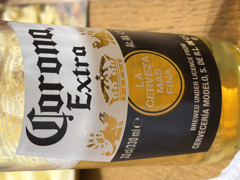 The image displays a clear glass bottle of Corona Extra beer, partially consumed, positioned centrally on a light brown, possibly wooden, surface. The white and black label is prominent, featuring the "Corona Extra" logo in black gothic script and standard serif font. Below this, gold illustrations of two camels and a crown are visible. A gold circular emblem with the text "LA CERVEZA MAS FINA" is centered below the crown. Additional text includes "33 cl / 330 ml e" on the left and "Alc. 4.5%" on the right, along with "BREWED UNDER LICENCE FROM CERVECERÍA MODELO, S. DE R.L. DE C.V." at the bottom. The bottle contains a light yellow liquid, with a visible line indicating partial consumption and residual foam bubbles along the interior glass near the top. The background is out of focus, suggesting an indoor setting. No individuals or specific actions are depicted. The image does not contain information to determine a specific location within Amber Valley, United Kingdom.