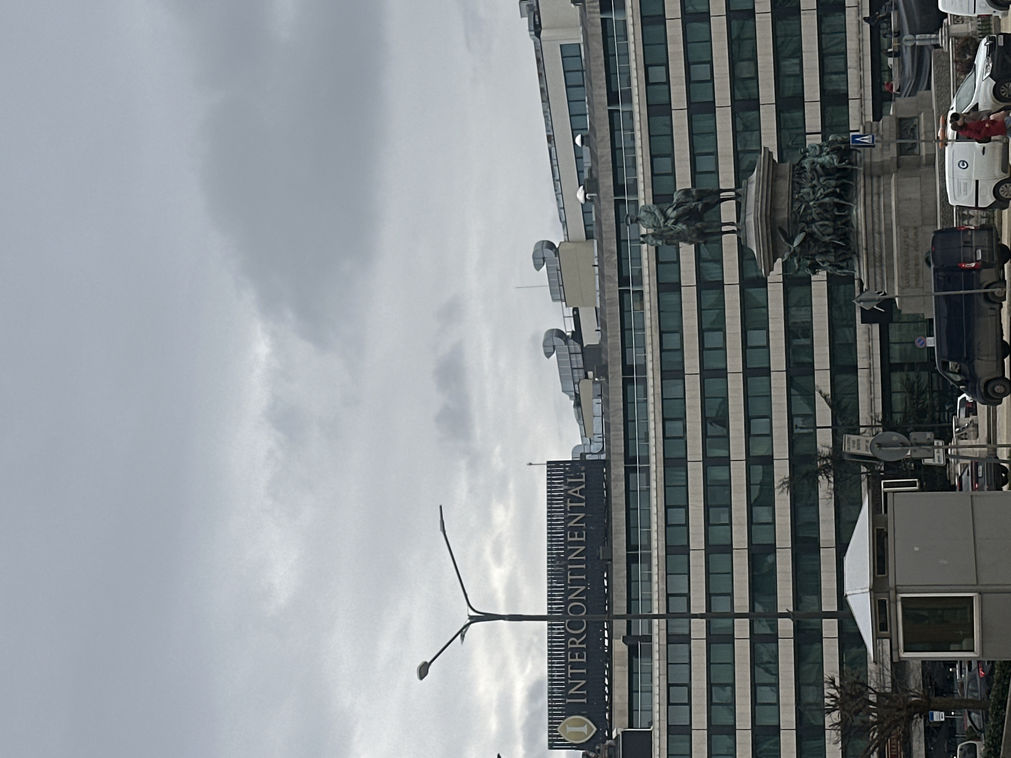 The image shows a view of Sofia, Bulgaria. The foreground features a sign for the InterContinental Hotel, with a tall building behind it. Adjacent to the hotel, a statue of a horse and rider, possibly a historical figure, is visible. Several cars are parked along the street. The sky is overcast.