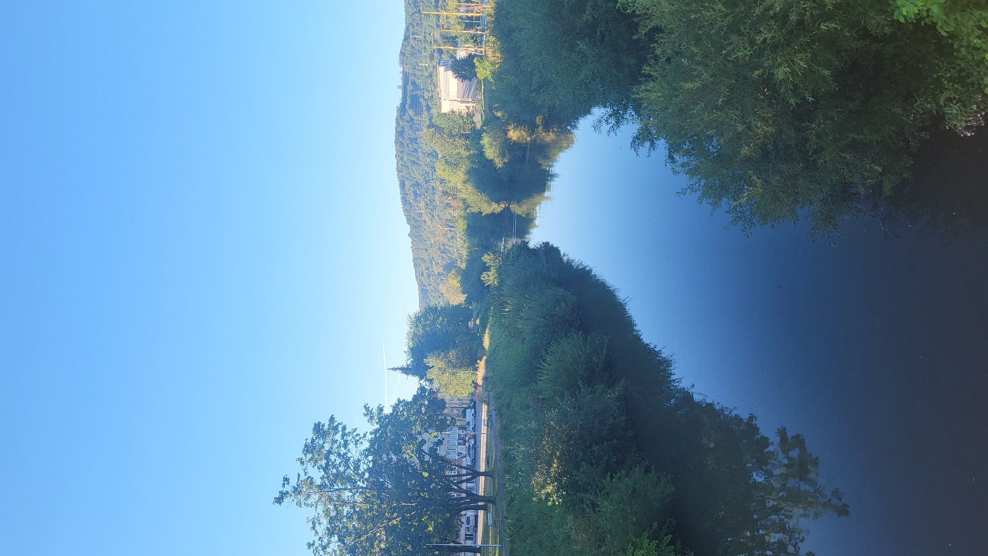 The image captures a tranquil scene of the Torysa River flowing through the city of Prešov, Slovakia. The river's surface reflects the clear blue sky, creating a sense of serenity. Lush green vegetation lines the riverbanks, with trees and bushes forming a natural border. In the background, a gentle hill covered in trees slopes downwards, suggesting a suburban or semi-rural setting within the city. The absence of people or any active human presence contributes to the peaceful ambiance of the photograph. The lighting indicates it is likely daytime, with the sun casting a soft glow on the landscape.