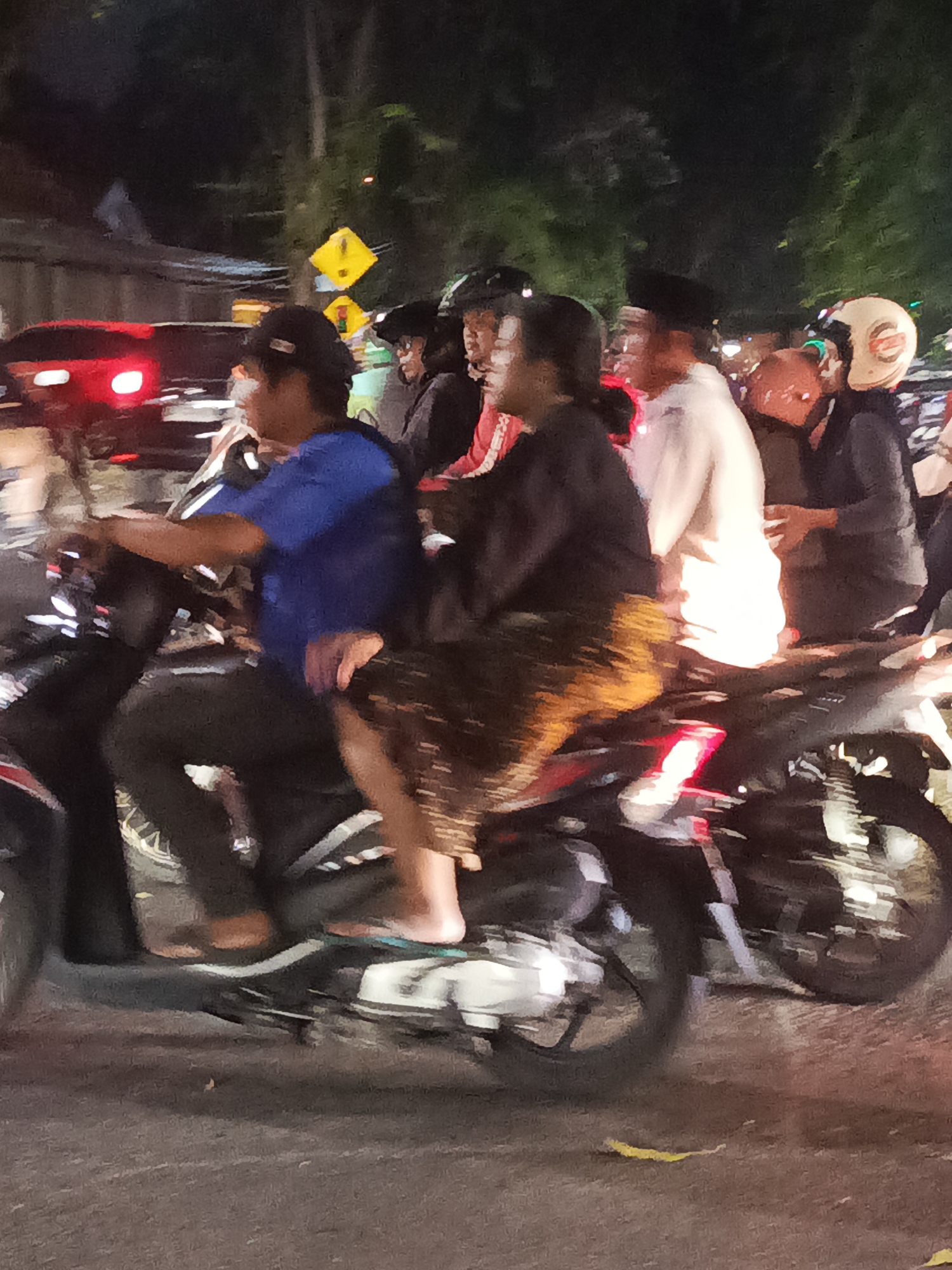 This is a nighttime, motion-blurred photograph taken on a street in Surabaya, Indonesia. The image captures several people on motorcycles, suggesting a busy urban environment.
In the foreground, two individuals are riding a motorcycle. The rider in front is wearing a blue shirt and dark pants, with a black baseball cap. They are operating the motorcycle. The passenger behind them is wearing a dark top and a patterned skirt or sarong, with their legs visible. Their left foot is bare, and their right foot is wearing a flip-flop.
Behind this motorcycle, other motorcycles and individuals are visible, also in motion. One person is wearing a white shirt and a black cap, possibly a peci, commonly worn in Indonesia. Another individual is wearing a beige helmet with an orange stripe. A red taillight from a vehicle is also visible in the background, indicating traffic.
The environment appears to be a paved street at night, with some trees and foliage visible in the background, partially illuminated by streetlights or vehicle lights. A yellow traffic sign with a black symbol is present on the left side of the image. The overall impression is of a dynamic street scene with multiple motorcycles navigating the road at night.