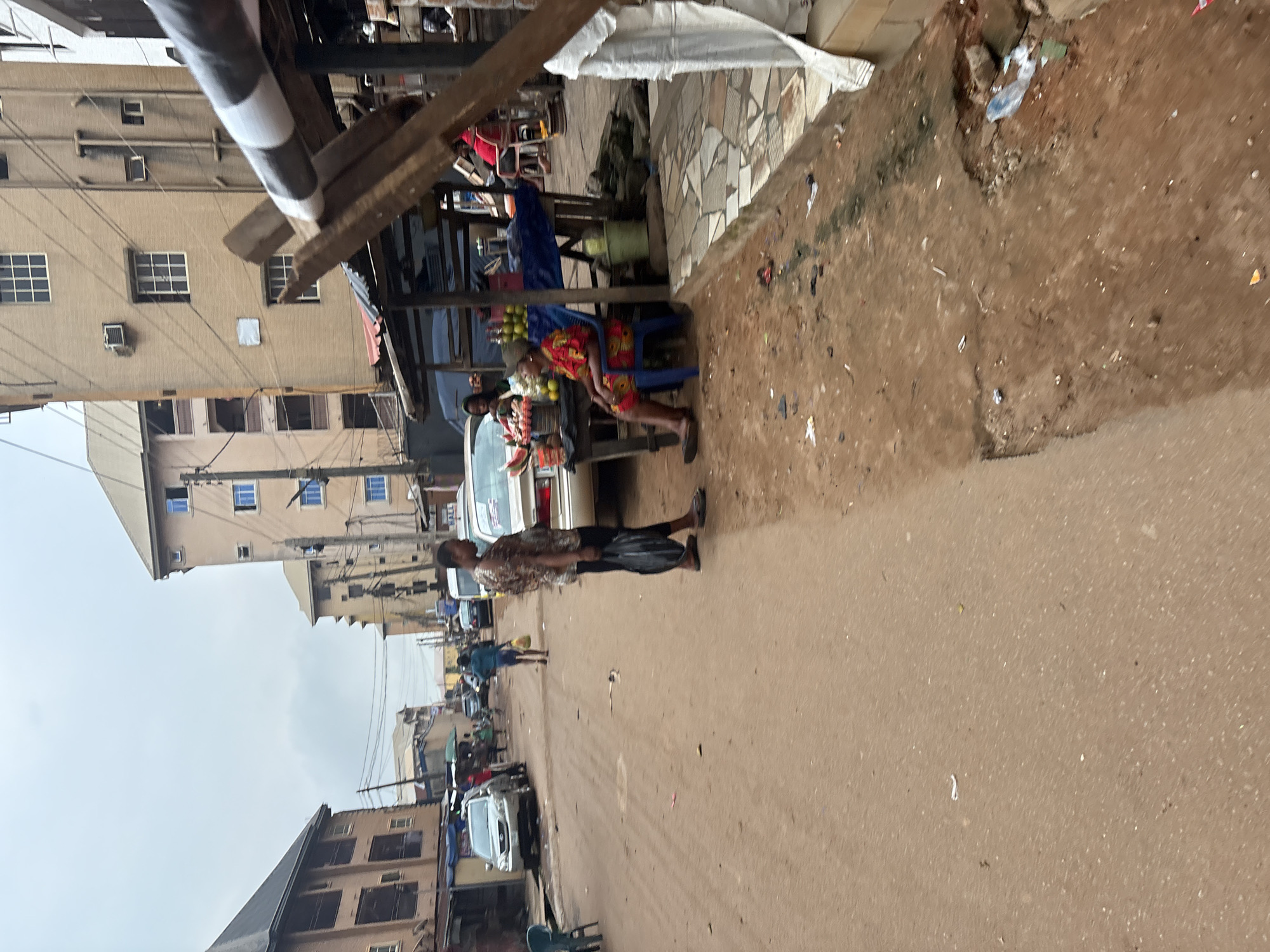 The image depicts a street scene in Onitsha, Nigeria, with a focus on a roadside market. In the foreground, a woman in a patterned dress is seated on a blue plastic chair at a market stall, which displays fruits and vegetables. A man in a patterned shirt and dark pants stands to her left, holding a black plastic bag. To the left of the market stall, a silver car is parked. Further down the street, several buildings line the road, some with multiple stories and balconies. A person in a blue shirt and dark pants walks away from the camera, carrying a yellow bag. In the background, more cars and people are visible, and power lines crisscross above the street. The ground is a mix of dirt and patchy pavement, with some debris present. The sky is overcast.