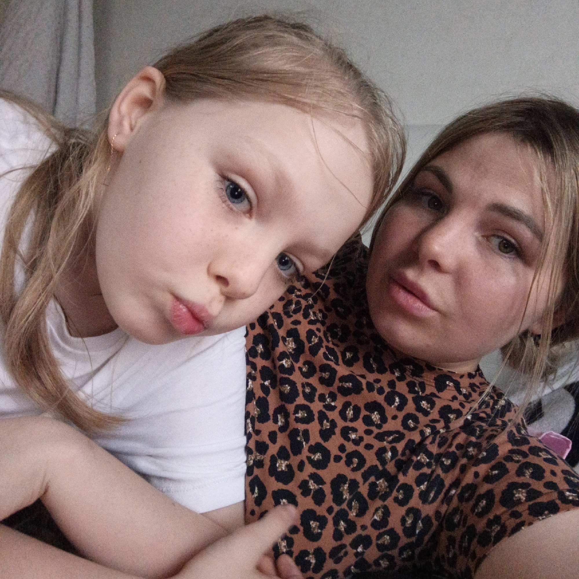 Two Caucasian females pose closely together indoors. The female on the left, likely a child, has fair skin, blue eyes, and light blonde hair. She wears a white T-shirt and has a small gold hoop earring. The female on the right has light brown hair and wears a brown and black leopard print top. The child rests her head on the adult’s shoulder, and both are pouting at the camera. The location is unknown.