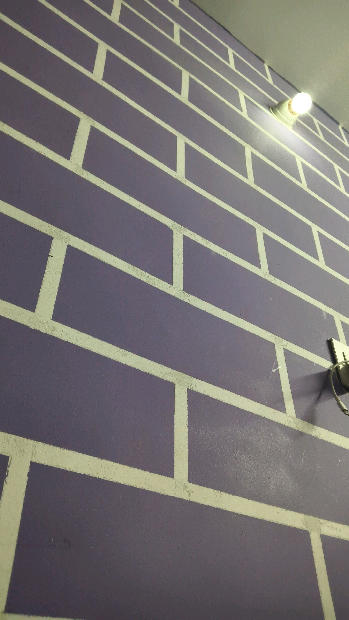 The image displays a close-up, angled view of a purple wall with white painted lines simulating a brick pattern. The lighting suggests an indoor environment, possibly a room. A lit light bulb in a white fixture protrudes from the wall near the top right, casting a warm glow. To the right of the light fixture, a white electrical outlet is visible with a cable plugged in and hanging down. The paint on the wall shows some texture, and the white lines have a slightly rough, uneven application. The perspective creates a sense of depth, drawing the viewer's eye up and along the wall. The exact location within Nandigama, India, is not determinable from the visual information provided.