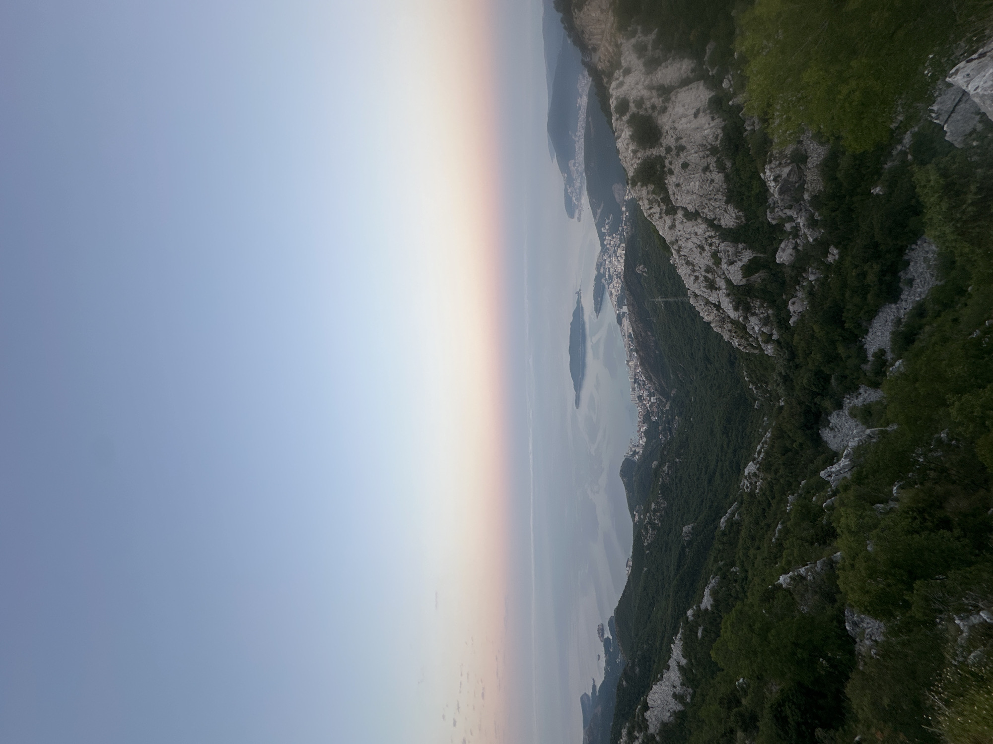 The image captures a panoramic view of Brajici, Montenegro, during what appears to be either sunrise or sunset. The sky transitions from a pale blue at the top to soft pink and orange hues near the horizon. Below the sky, the Adriatic Sea stretches out, calm and reflecting the sky's colors. A town with white buildings is visible along the coastline, nestled between the sea and the rugged, green mountains. The foreground consists of rocky, mountainous terrain covered with dense green vegetation. There are no people or animals visible in the image. The overall atmosphere is serene and offers a sweeping vista of the coastal landscape.