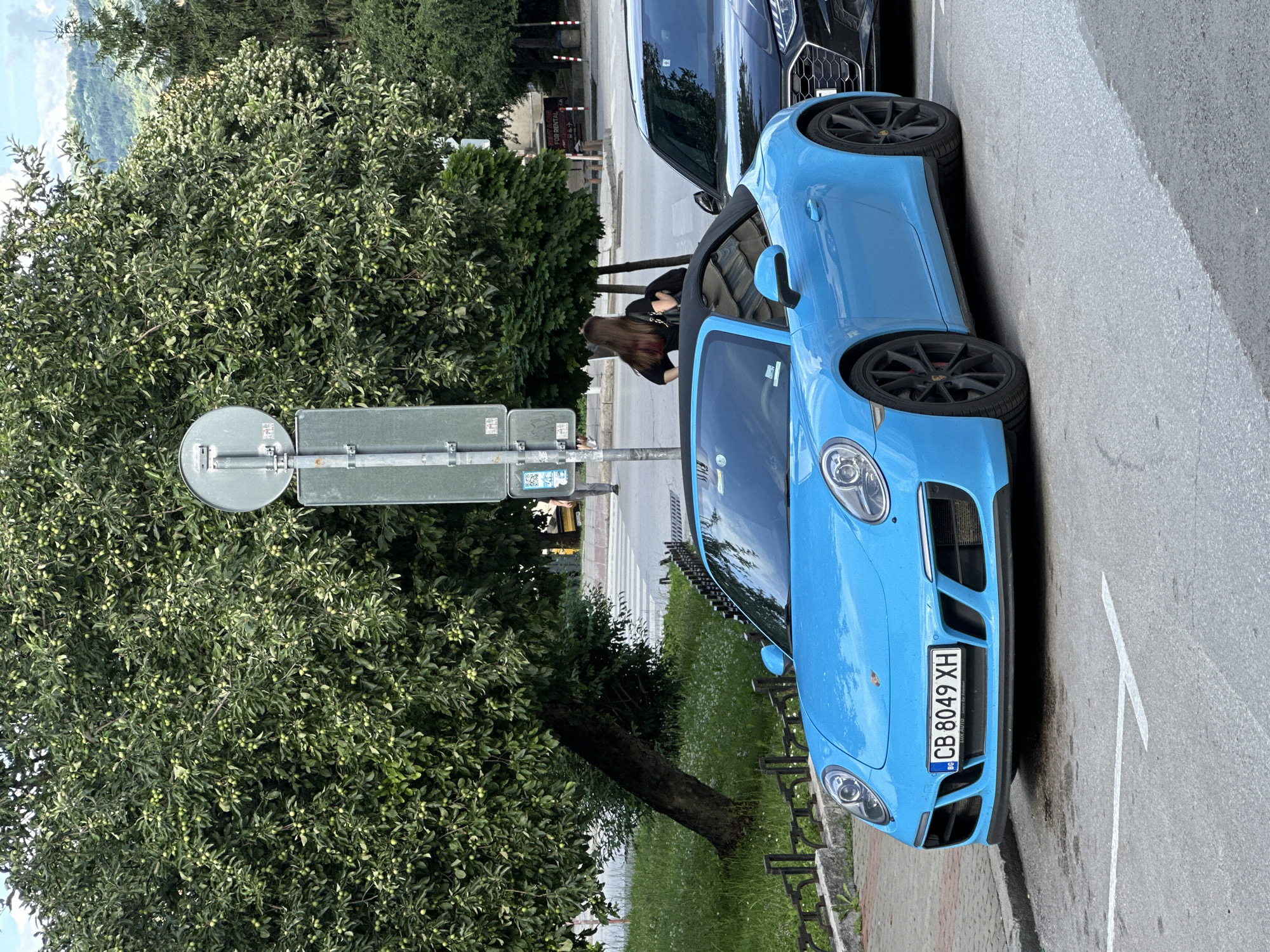 A bright blue convertible Porsche is parked on a city street in Veliko Tarnovo, Bulgaria, next to a dark-colored sedan. A woman with long brown hair, wearing a black top and carrying a black handbag, stands behind the Porsche, facing away from the camera. She appears to be looking towards a pedestrian crossing. The street is paved, and there are parking lines visible. Behind the cars, there is a grassy area with a leaning tree that has green foliage. In the background, a street sign with multiple panels is visible, as well as more trees and a hint of a hillside. The sky is overcast.
