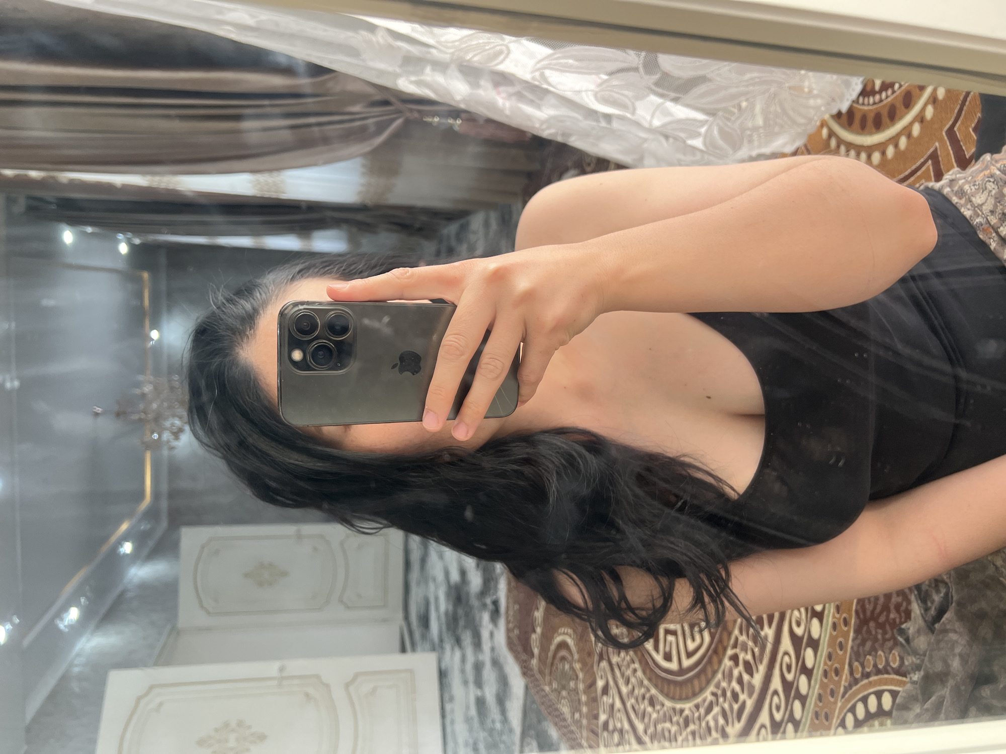 The image shows a woman taking a mirror selfie. She has long, dark hair and is wearing a black tank top. She is holding a smartphone in her right hand, which is in front of her face. The background is a reflection of a room, which appears to be a bedroom. There is a large rug on the floor, with an ornate pattern. There are also curtains and doors visible in the reflection. The overall impression is one of a casual, domestic setting. The provided location of Kara-Dıykan, Kyrgyzstan, does not offer any specific visual cues within the image to confirm or deny its presence.