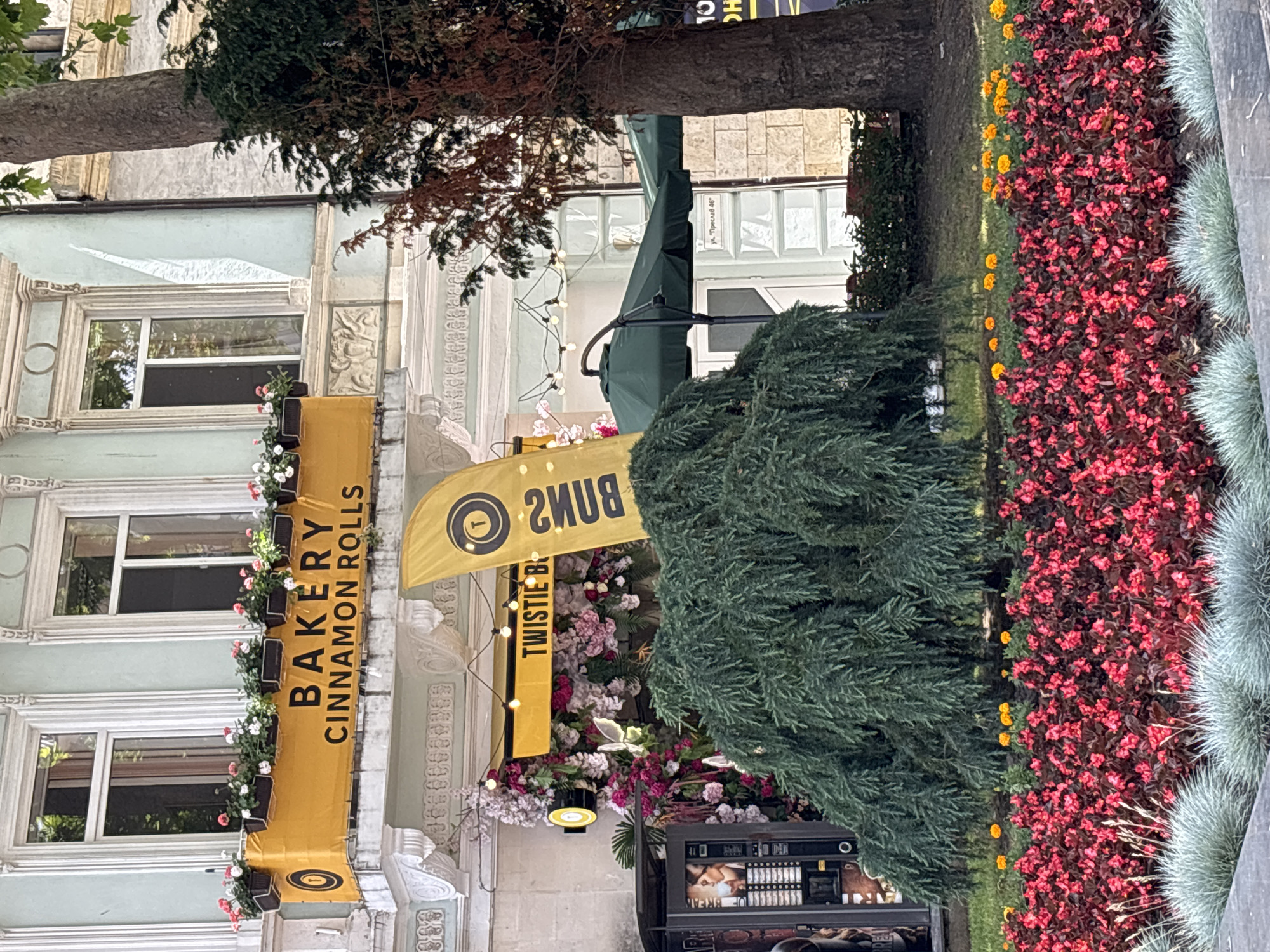 The image depicts a street-level view of a bakery in Varna, Bulgaria, specifically located on Preslav Street 46. A prominent yellow banner above the shop entrance reads "BAKERY CINNAMON ROLLS". Another yellow flag to the right, with a spiral logo, has text that appears to be "TWISTIE B..." and then possibly "...BAKERY". The shop facade is decorated with abundant floral arrangements in shades of pink and white, with fairy lights strung above. To the left of the entrance, a vending machine is visible. In front of the bakery, a large, weeping evergreen tree partially obscures a seating area with green umbrellas. A vibrant flower bed with red and pink blossoms, interspersed with yellow marigolds, occupies the foreground. Several people are not visible in the image.
