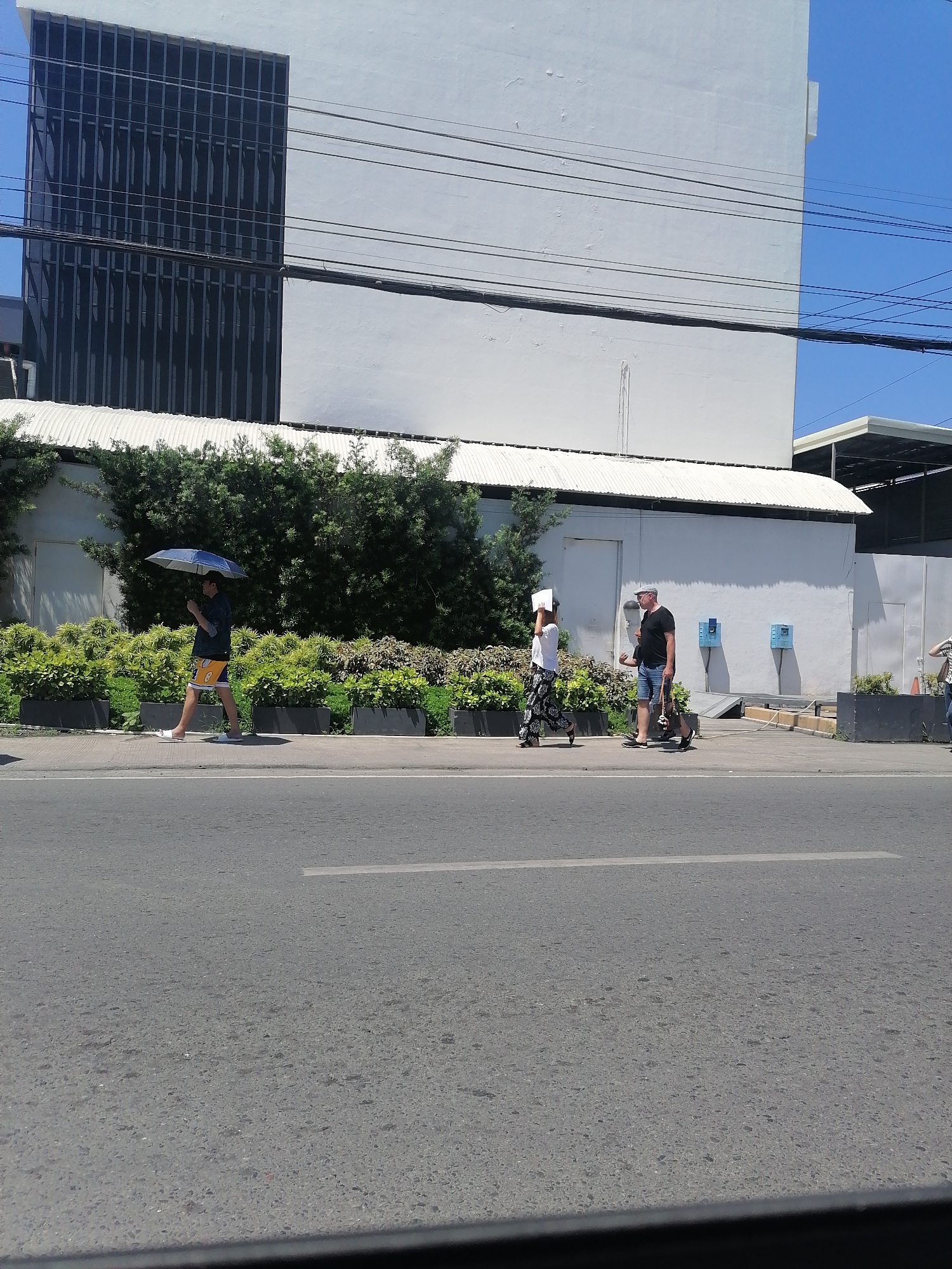 A street scene in Davao City, Philippines shows three people walking on the sidewalk in front of a white building with a metal roof and a dark-paneled section on the left. The first person, on the far left, is a man with dark hair walking under a blue umbrella. He is wearing a dark shirt and yellow shorts with white stripes. The second person, in the middle, is a woman wearing a white t-shirt and black and white patterned pants, carrying a white piece of paper. She is walking next to a dark-colored stroller. The third person, on the right, is a man wearing a black t-shirt and blue shorts, with a gray cap. He is walking with a woman behind him who is wearing a white shirt and blue jeans and has light-colored hair. The sidewalk is lined with manicured green plants in dark rectangular planters. A paved road with a white lane marker runs in front of the sidewalk. Power lines are visible in the sky above the building.