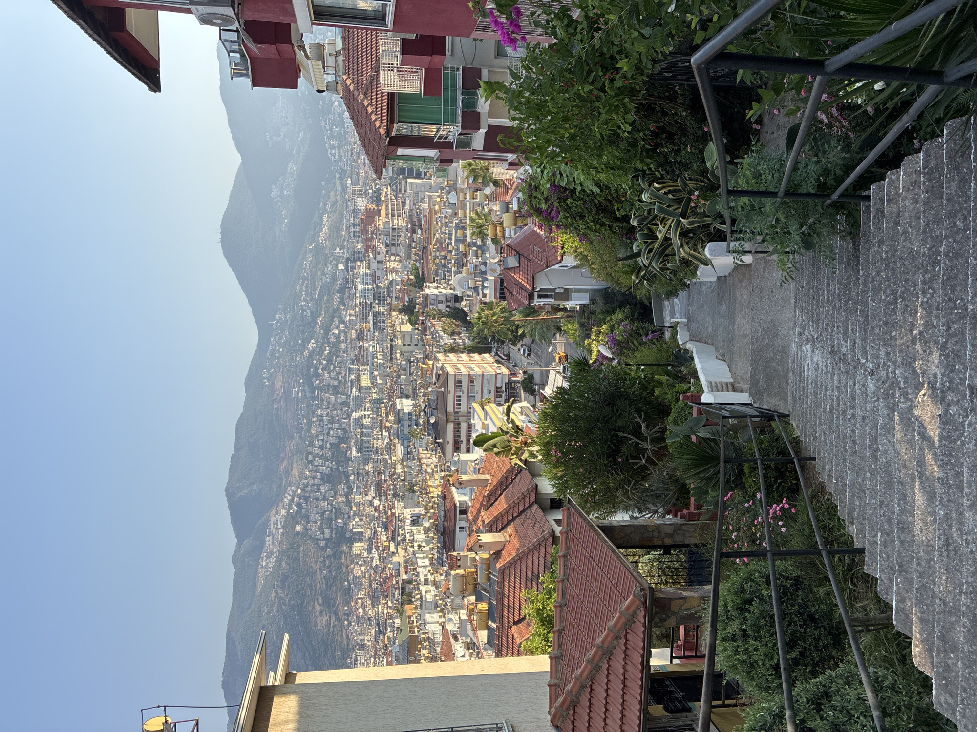 The image depicts a downward view of a stone staircase in Alanya, Turkey, flanked by lush greenery and railings. The staircase leads towards a densely populated city below, characterized by numerous buildings with red-tiled roofs and solar water heaters. In the background, rolling hills covered in vegetation and scattered buildings rise towards a clear, pale blue sky. The scene appears to be captured in the early morning or late afternoon, indicated by the soft sunlight illuminating the structures and foliage. The architecture suggests a Mediterranean or similar climate, with the prominent use of solar water heating systems being common in the region. No people are visible in the frame.