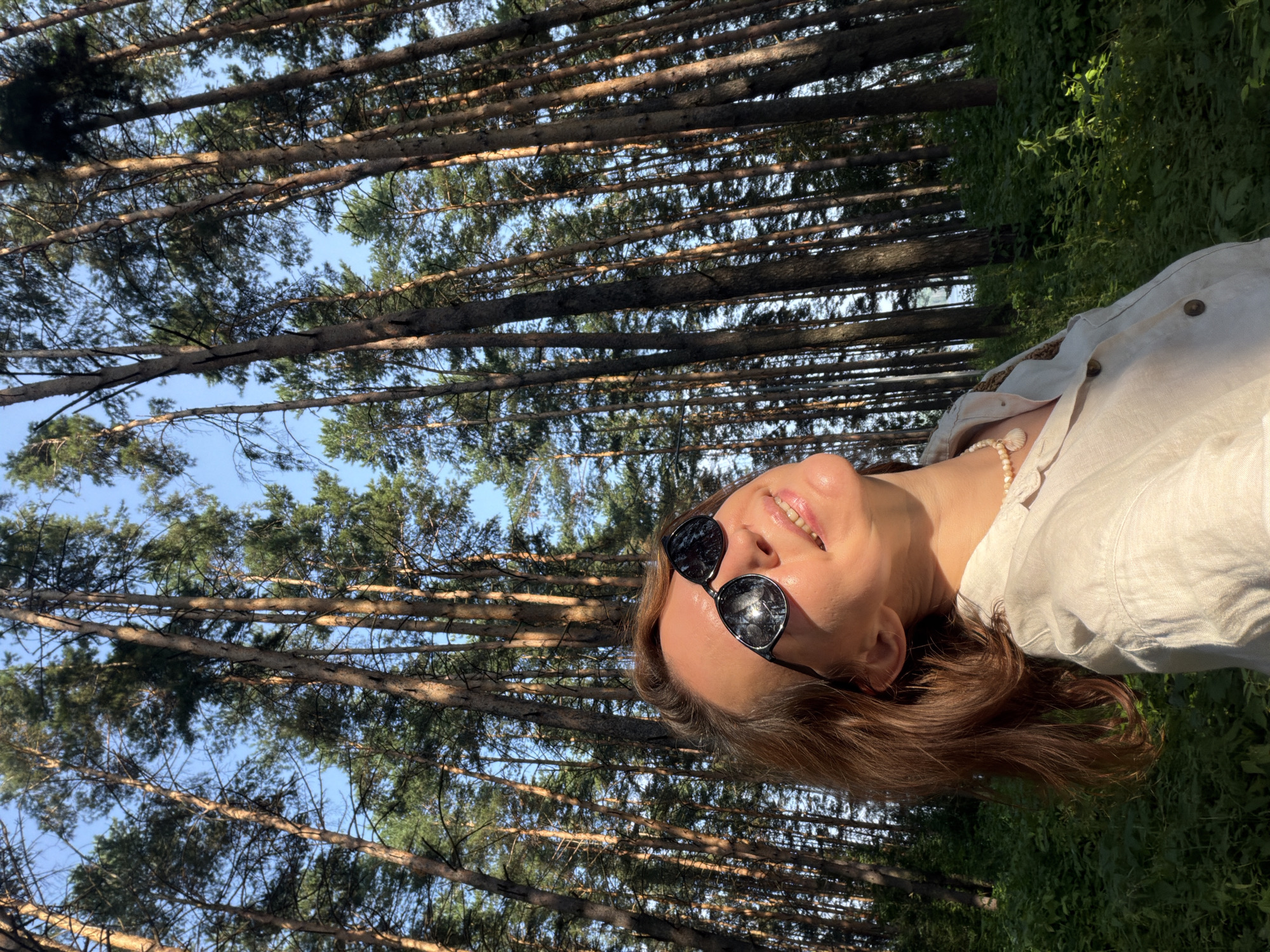 A low-angle, medium shot captures a Caucasian woman outdoors in a forest. The woman, with reddish-brown hair, wears large, dark sunglasses and a light-colored, collared shirt. A pearl necklace with a shell pendant is visible around her neck. She is looking upwards and to her right, with her head tilted back and her mouth slightly open in a smile. The background is dominated by tall, slender pine trees reaching towards a clear blue sky, with lush green foliage visible at the base of the trees. The lighting suggests it is daytime, with sunlight casting shadows and highlighting the woman's face and the tree trunks. The image appears to be taken in the city of Chaikovsky, Russia, possibly in a park or natural area featuring a pine forest.