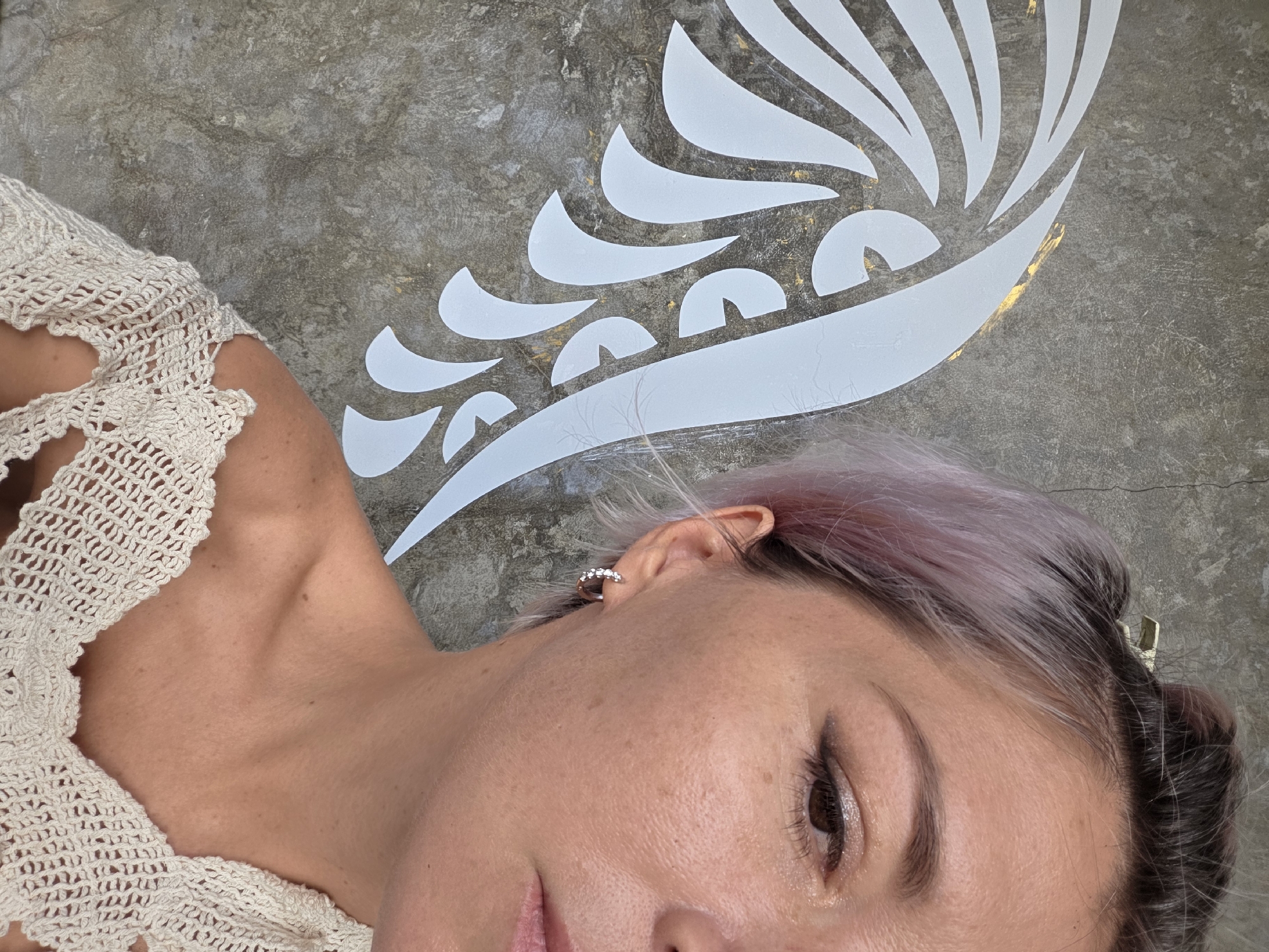 A close-up portrait of a woman is shown in North Kuta, Indonesia. The woman, with short, pink-tinted hair pulled back with a clip, is positioned to the right of the frame, with only the right side of her face visible. She is looking down and to her right. She is wearing a cream-colored, crocheted top that exposes her shoulders and upper chest. A diamond hoop earring adorns her left ear. To the left of the woman, a large, white, stylized wing design is painted on a textured concrete wall. The wall itself has a gray, industrial appearance, with some subtle gold accents visible behind the wing. The lighting suggests an outdoor setting, possibly during the day.
