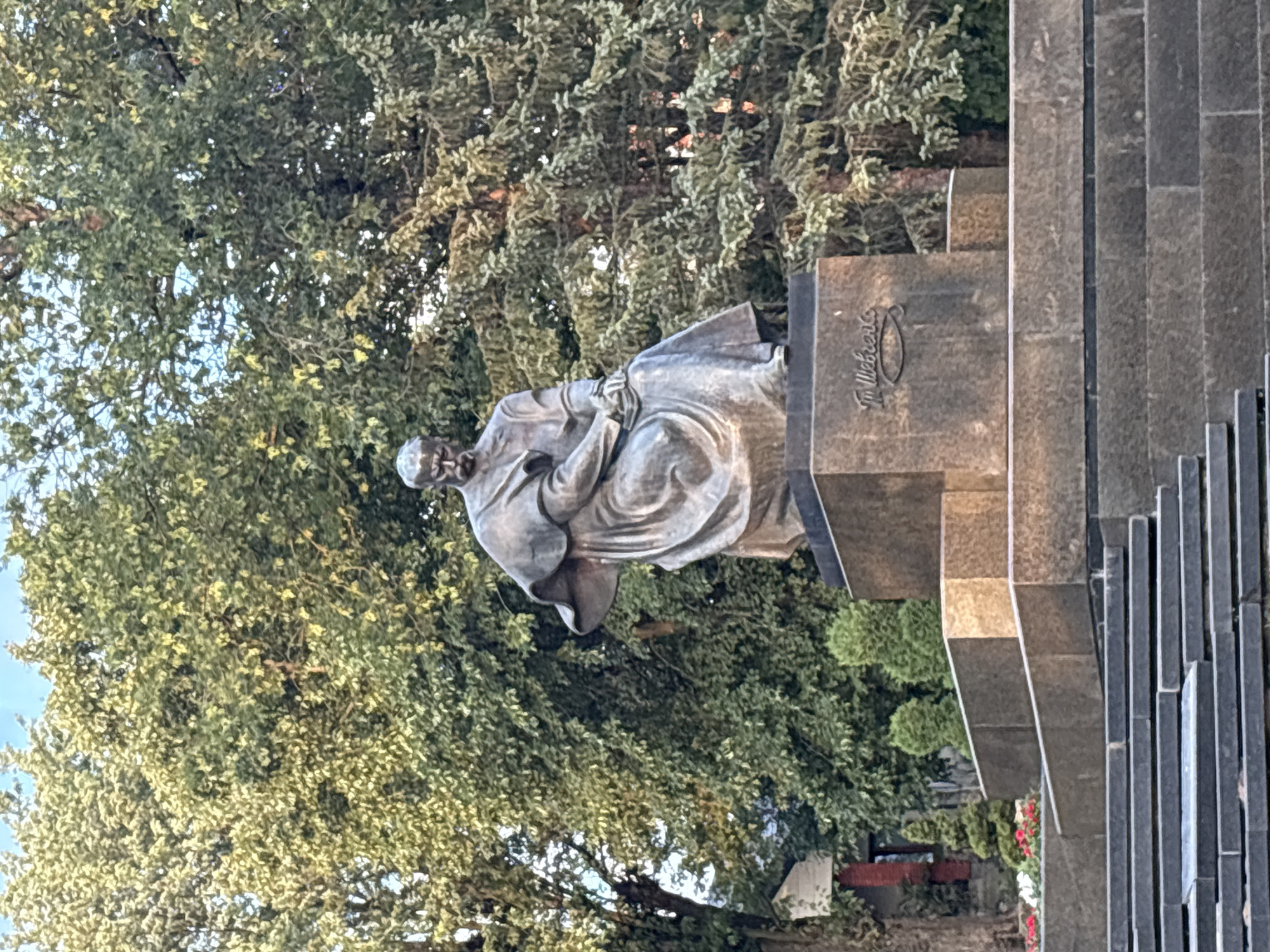 A bronze statue of a seated man is positioned on a granite pedestal in front of a dense background of green trees. The man wears a robe and has his arms resting on his lap. The pedestal has an inscription. The statue is located in the city of Kalush, Ukraine, specifically in Shevchenko Square, dedicated to the Ukrainian national poet Taras Shevchenko. The foreground consists of a wide stone staircase leading up to the monument.