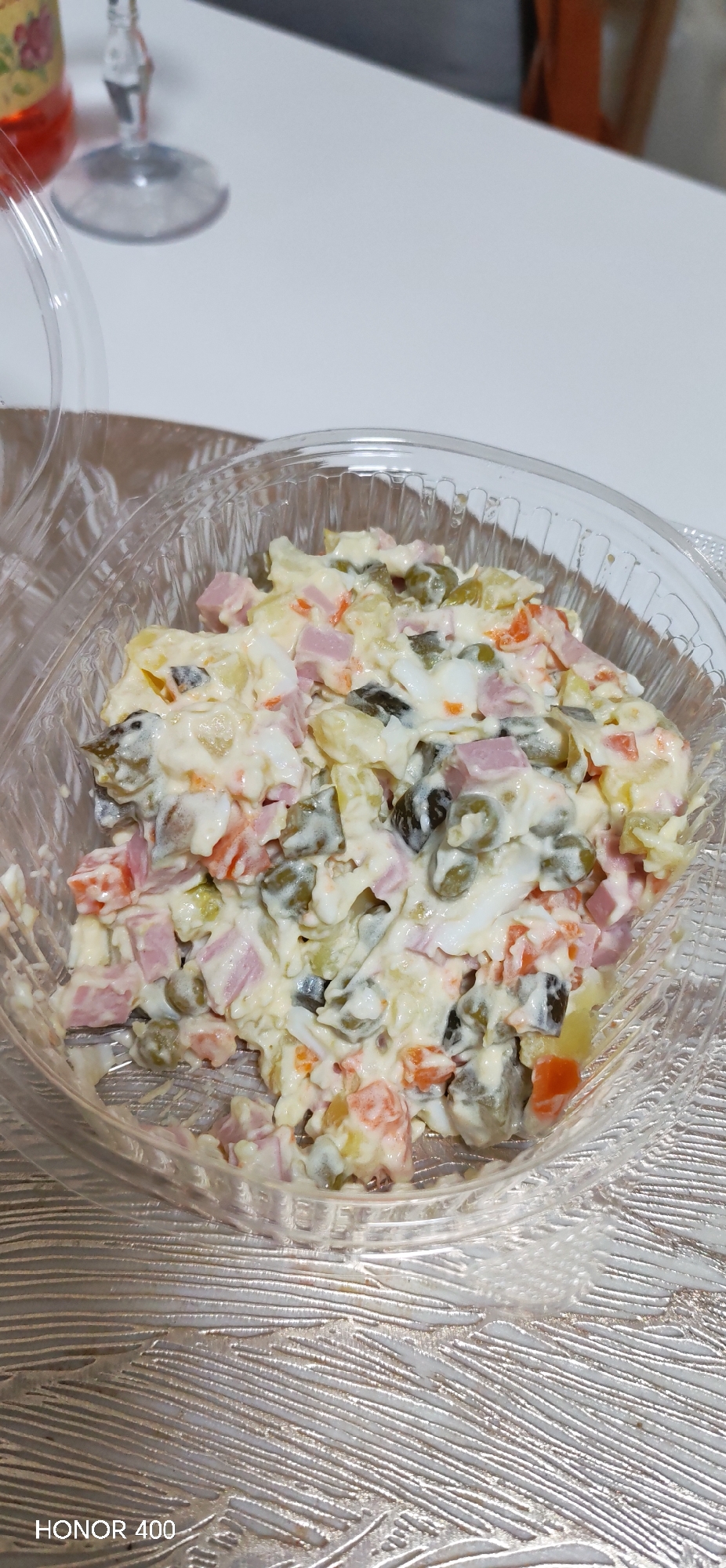 A clear plastic bowl filled with Olivier salad sits on a textured, reflective surface. The salad contains diced potatoes, carrots, peas, pickles, ham, and mayonnaise. A glass with a beverage is visible in the background. The inscription "HONOR 400" is at the bottom left of the image. There are no apparent indicators of a location in Noviy, Russia.