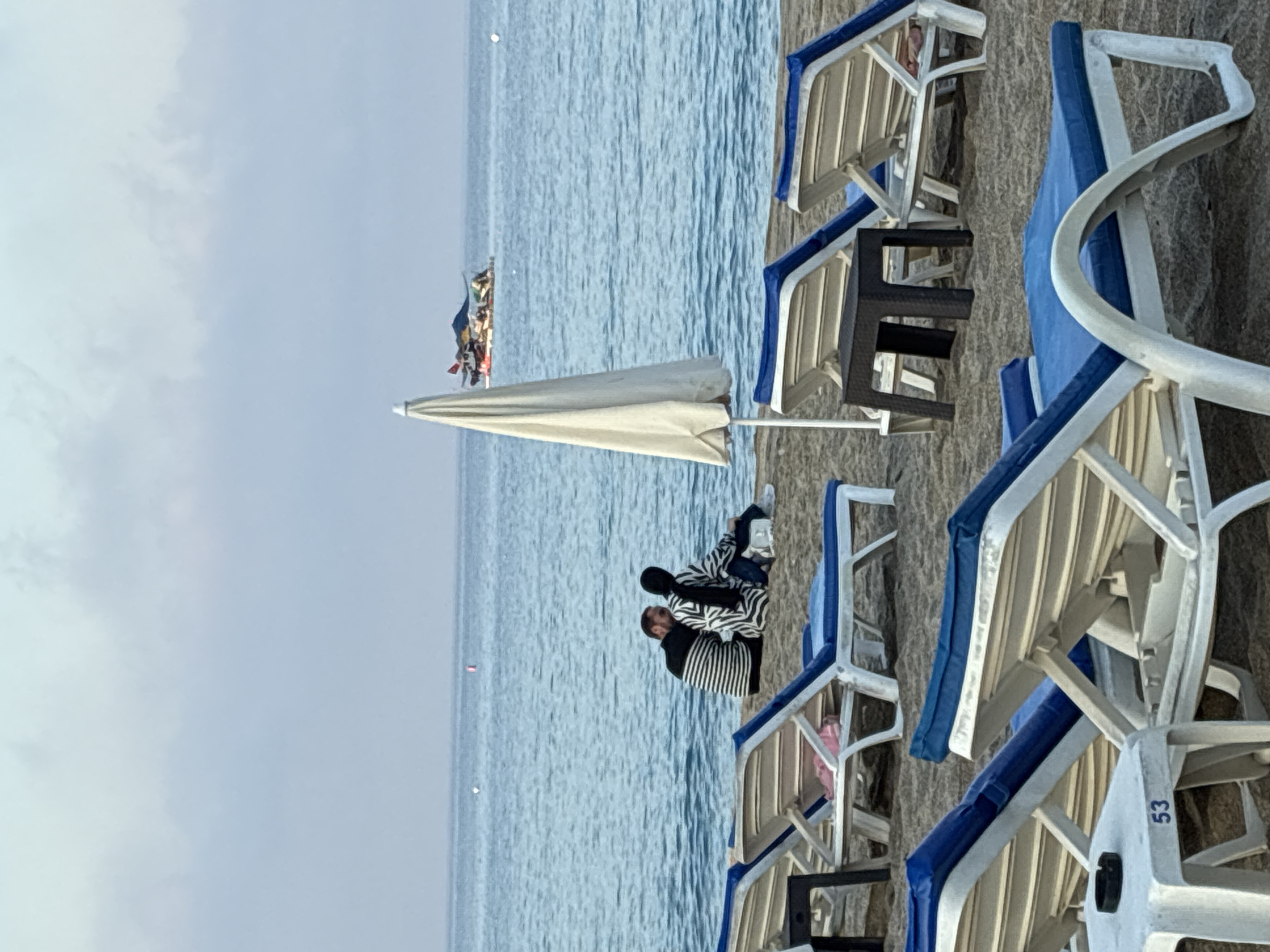 A couple is sitting on a beach in Alanya, Turkey, looking out at the calm Mediterranean Sea. The man is wearing a striped shirt, and the woman is wearing a headscarf and a patterned top. They are seated on the sand, with several empty lounge chairs in the foreground. In the background, a small boat is visible on the water. The sky is overcast, and the overall atmosphere is peaceful.