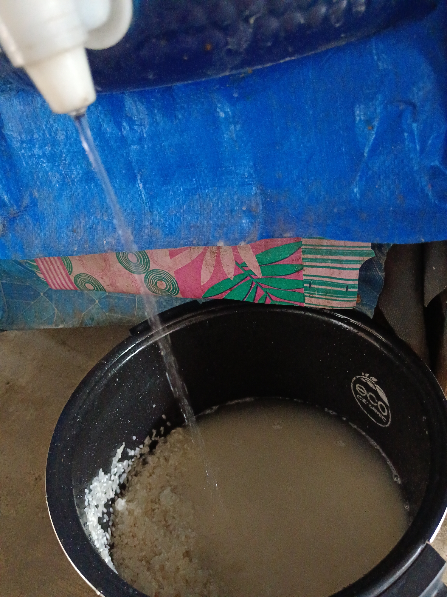 In a residential setting, possibly a kitchen, in Aek Kanopan, Indonesia, water is being poured from a white dispenser into a black rice cooker. The rice cooker contains uncooked rice and water. Behind the rice cooker, a blue tarp is visible, with a patterned fabric strip draped over it. The floor appears to be concrete. The action suggests the preparation of rice.