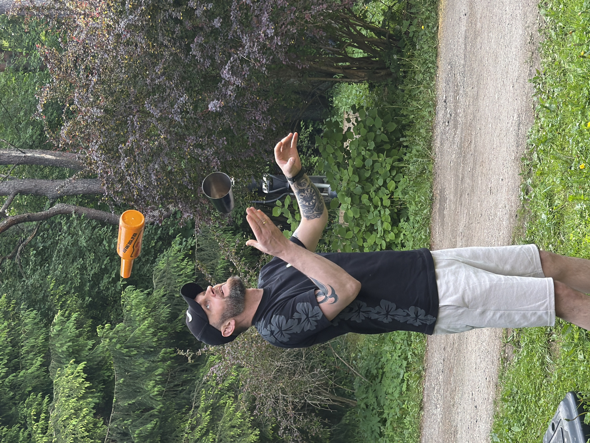 A man with a beard and tattoos on his arm is juggling an orange bottle and a metal cup in an outdoor setting with trees and bushes. He is wearing a black t-shirt with a floral pattern on the sleeves, a black baseball cap, and light-colored shorts. The man is looking up at the objects he is juggling. The environment suggests a park or a wooded area. The image is taken from a medium distance, capturing the man from the waist up. The lighting indicates it is daytime, with sunlight filtering through the trees. There is a gravel path in the foreground, and lush green foliage surrounds the scene.