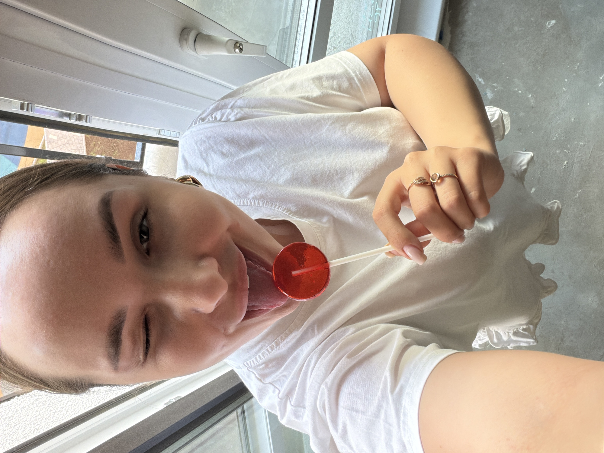 A young woman is close to a window inside a building in Pervomaysky, Russia. She is wearing a white t-shirt and has jewelry on her fingers. She is holding a red lollipop with a white stick and sticking her tongue out to lick the lollipop, while winking.