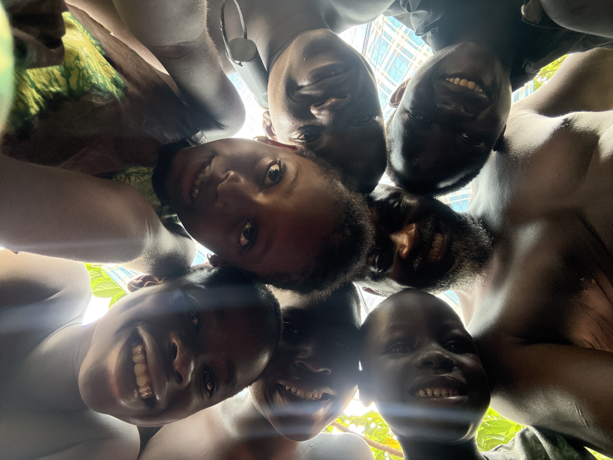 A low-angle, close-up, indoor shot captures a group of seven young Black individuals, their faces pressed together in a circle looking upwards towards the camera. The subjects are positioned in a tight cluster, with some faces partially obscured by others. Sunlight streams in from above, creating lens flares and highlighting the subjects' features. The individuals appear to be smiling and engaged. One individual in the foreground, wearing a dark shirt, has a stethoscope draped around their neck. Another individual, with a beard, is visible in the upper left quadrant of the image. The background consists of a grid-like pattern that could be a window or a screened-in area, and glimpses of green foliage suggest an outdoor environment visible through this structure. The overall impression is a candid and joyous moment captured in what appears to be a residential setting in Ado, Nigeria.