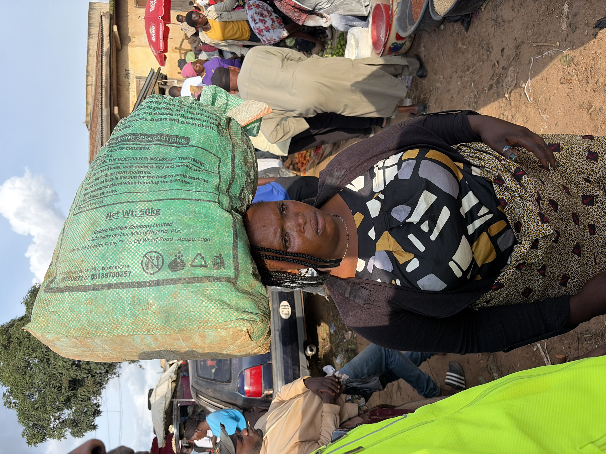In Gura Top, Nigeria, a woman with braided hair carries a large, green sack of grain on her head in a bustling outdoor market. She wears a patterned dress and a black cardigan. The sack displays text including "Golden Fertilizer Company Limited" and "Net Wt.: 50kg." In the background, people mill about, with stalls and a parked car visible. The ground is dirt, suggesting a typical market setting.