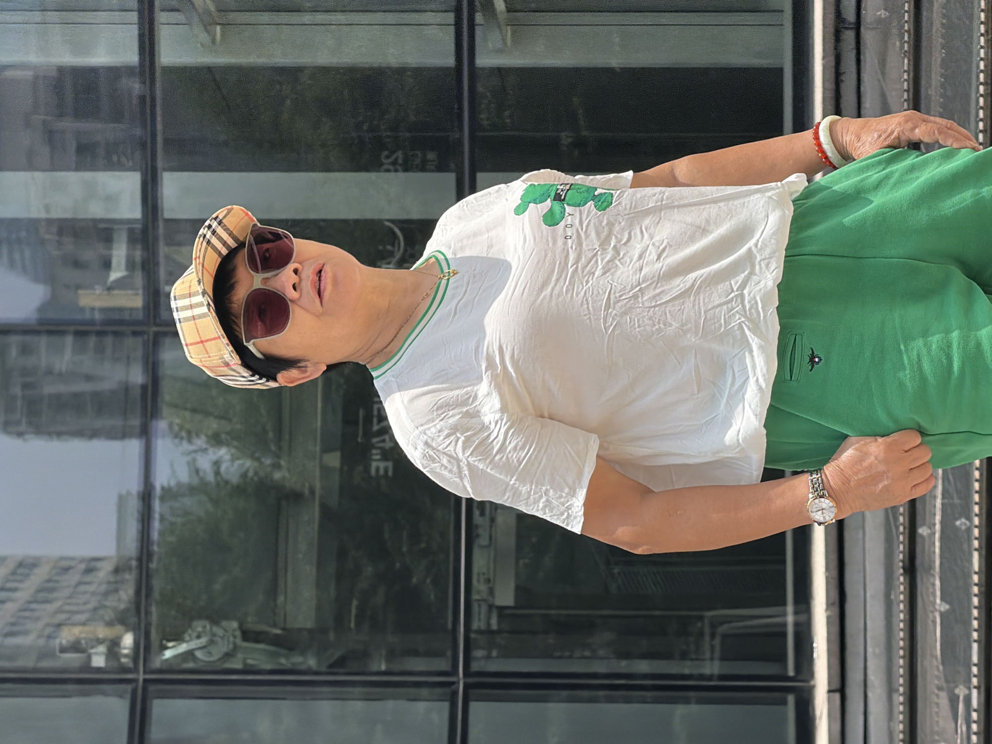 A person stands outdoors, in front of a modern building with large glass windows, likely in the city of Yuzhong District, China. The person is wearing a white t-shirt with a green trim around the collar and a graphic of a green cartoon bear on the left chest, a pair of green shorts, a plaid baseball cap, and sunglasses. They are also wearing a watch on their left wrist and two bracelets on their right wrist. The building's glass facade reflects the surrounding environment.