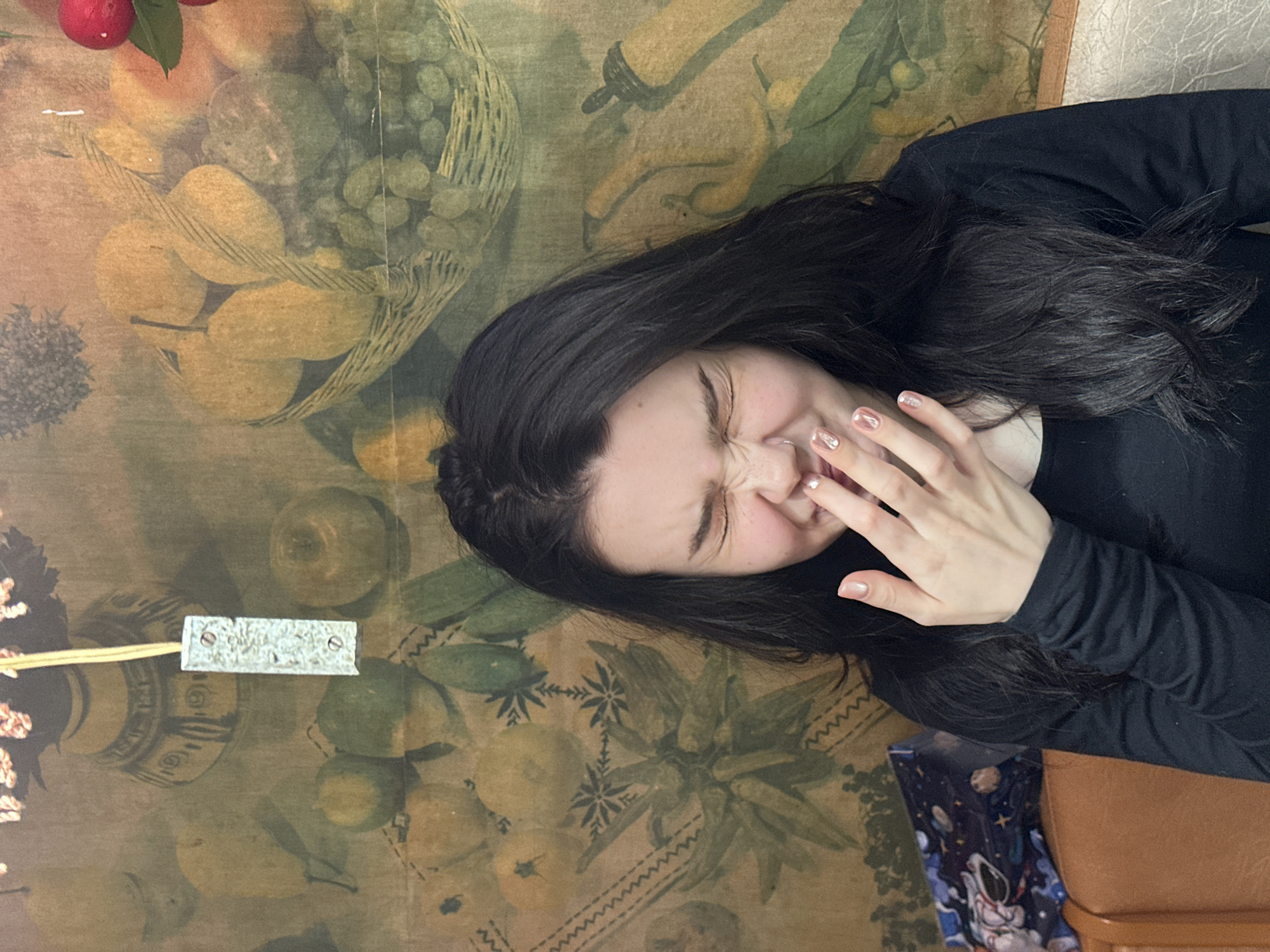 An individual with long dark hair, parted on the left, occupies the central foreground, wearing a black long-sleeved top. Their eyes are tightly closed, the nose is scrunched, and a small septum piercing is visible. Their left hand, displaying light, iridescent nail polish with a small white embellishment on one nail, covers the lower part of their face.
The background is a wall covering featuring a still-life pattern of fruits and vegetables in muted earthy tones, including pears, grapes in baskets, lemons, and other produce, interspersed with decorative elements such as zigzag lines and star-like motifs. A rectangular, metallic-colored object, possibly a switch or sensor, hangs from a thin yellow cord in the upper central portion of the frame. A small red fruit with green leaves is partially visible in the upper right corner.
To the lower left, a section of brown upholstered seating is visible, partially covered by a dark, patterned item featuring space-themed graphics. To the lower right, a lighter-colored, textured upholstered surface is present.
The scene is located in Aktanysh, Russia.