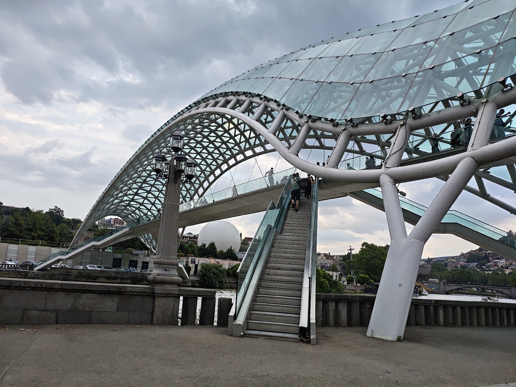 The image captures a view of the Bridge of Peace in Tbilisi, Georgia, a modern pedestrian bridge spanning the Kura River. The bridge features a distinctive glass and steel canopy that arches gracefully over the walkway. In the foreground, a set of outdoor stairs with glass railings leads up to the bridge. A few people are visible on the stairs and on the bridge itself. To the left, a large white domed structure, possibly an exhibition hall or museum, is visible on a hillside behind a stone embankment lined with cars. The background shows a cityscape with buildings and lush green trees, under a cloudy sky. The overall scene depicts a public space with architectural interest and urban activity.