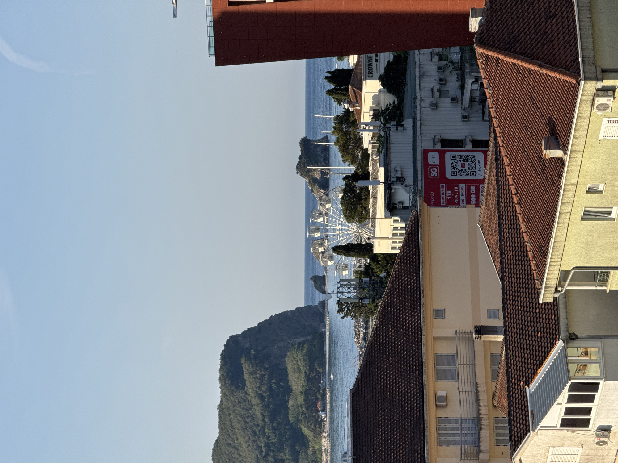 The image depicts a coastal city with a view of the sea and a large, verdant hill rising from the water on the left. Several buildings with red-tiled roofs dominate the foreground and midground. In the midground, a white Ferris wheel is visible, suggesting an amusement area or park. A tall, dark red building with modern architecture stands on the right side of the frame. The sky is clear and blue. The presence of the Ferris wheel and the coastal setting strongly indicate the location is Budva, Montenegro, a popular tourist destination known for its beaches and historic old town, often featuring such scenic views.