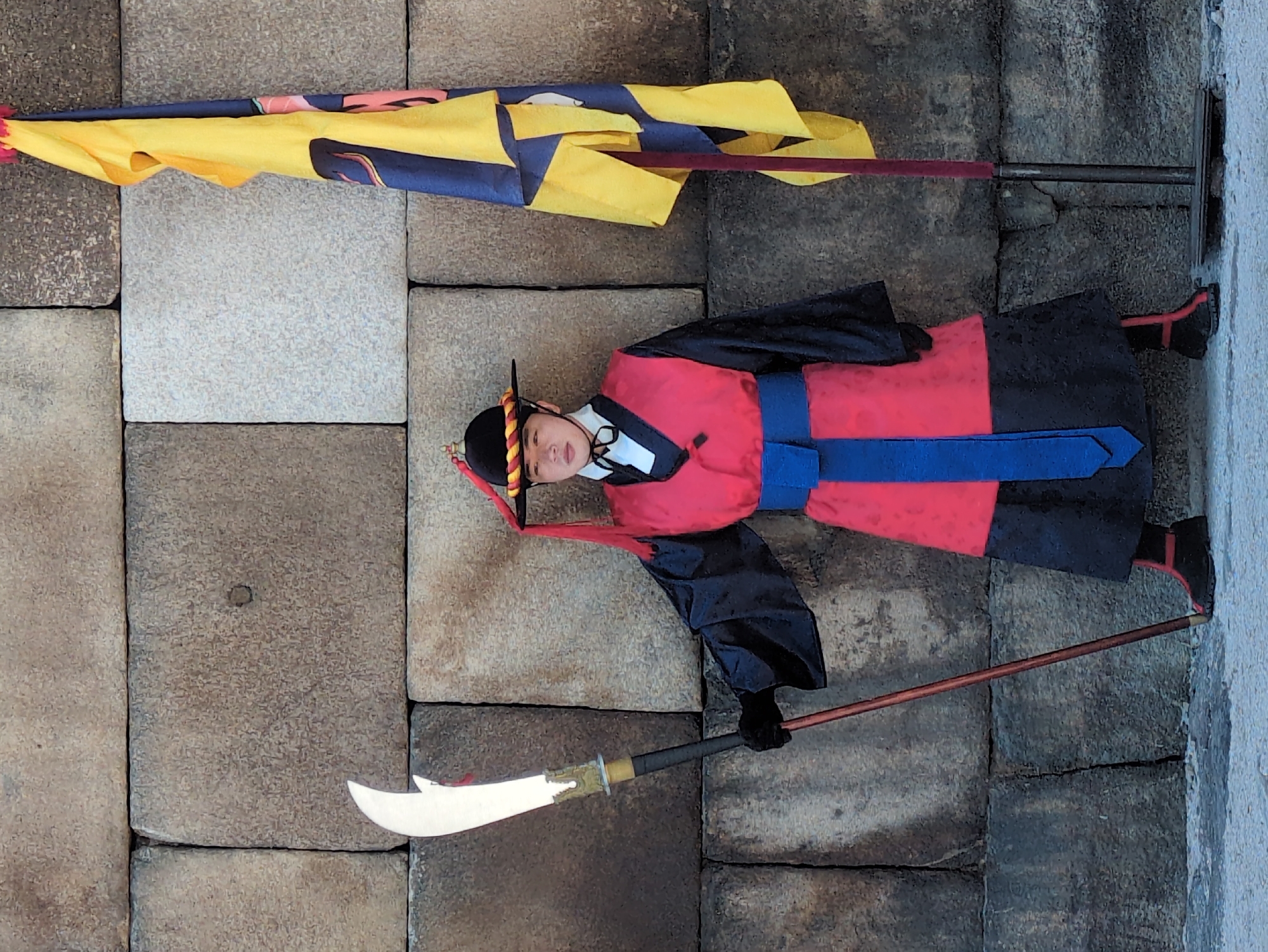 An overhead shot depicts a guard at Gyeongbokgung Palace in Seoul, South Korea. The guard is standing on a stone wall, dressed in a traditional red and black uniform with a blue sash. He wears a black hat with red tassels. The guard is holding a long-handled blade in his right hand and standing in front of a yellow and blue flag. The environment is a stone wall of the Gyeongbokgung Palace.