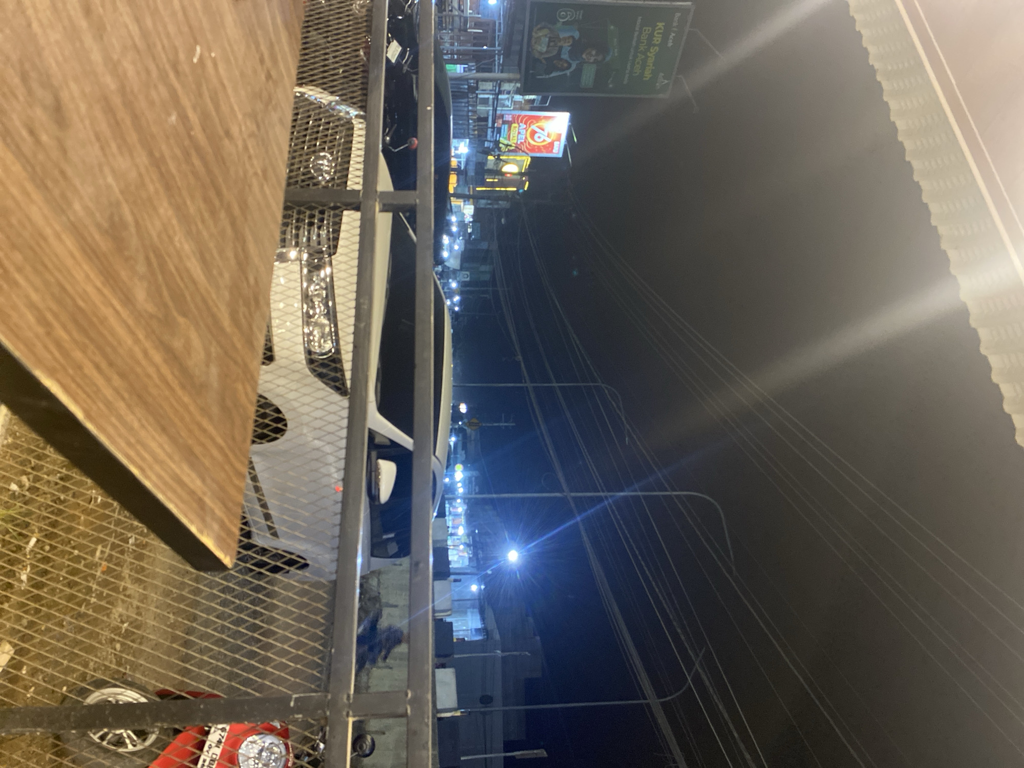 The image depicts a nighttime scene in Tanjongkapai, Indonesia. A wooden table is visible in the foreground, along with a metal structure resembling a fence or railing. A white car is parked on the street. In the background, there is a street with various signs, including one for "KUR Syariah Bank Aceh" and another for "76". Streetlights illuminate the scene, and power lines stretch across the dark sky. A red object with a headlight is visible below the fence, possibly a scooter.