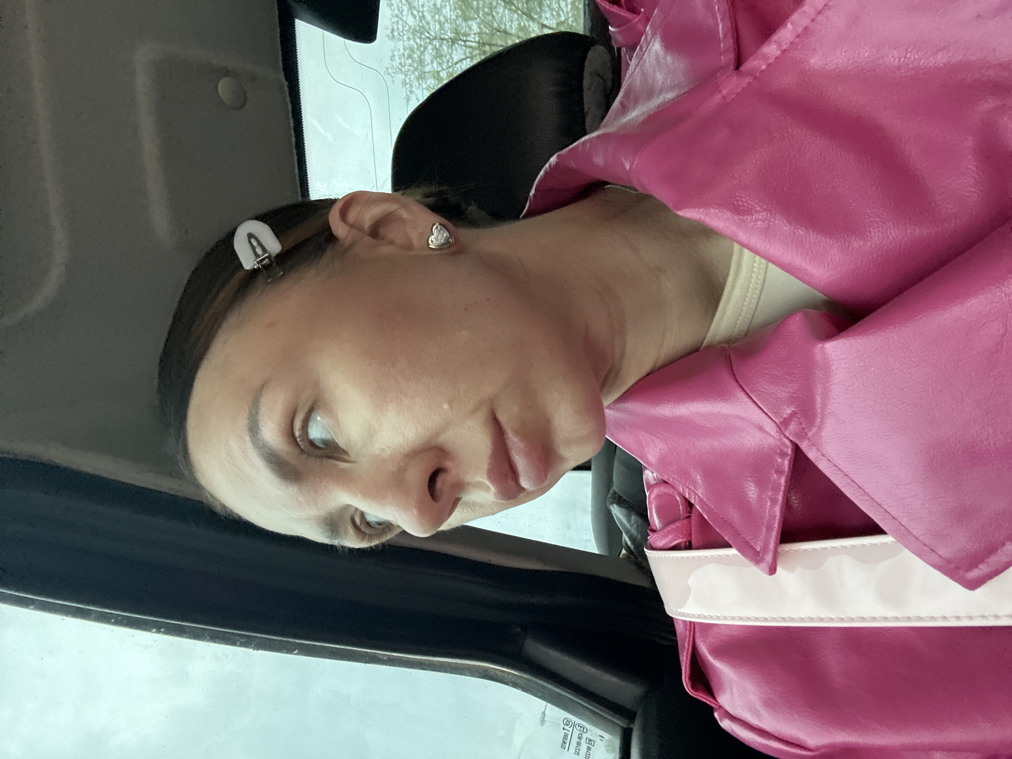 A woman is inside a vehicle, possibly a car, looking towards the camera with a slightly inquisitive expression. She is wearing a bright pink, possibly leather, jacket. A white clip is visible in her hair, and a heart-shaped earring adorns her ear. The background appears to show a window and hints of outdoor greenery. There is no specific indication in the image to confirm the location in Votkinsk, Russia.